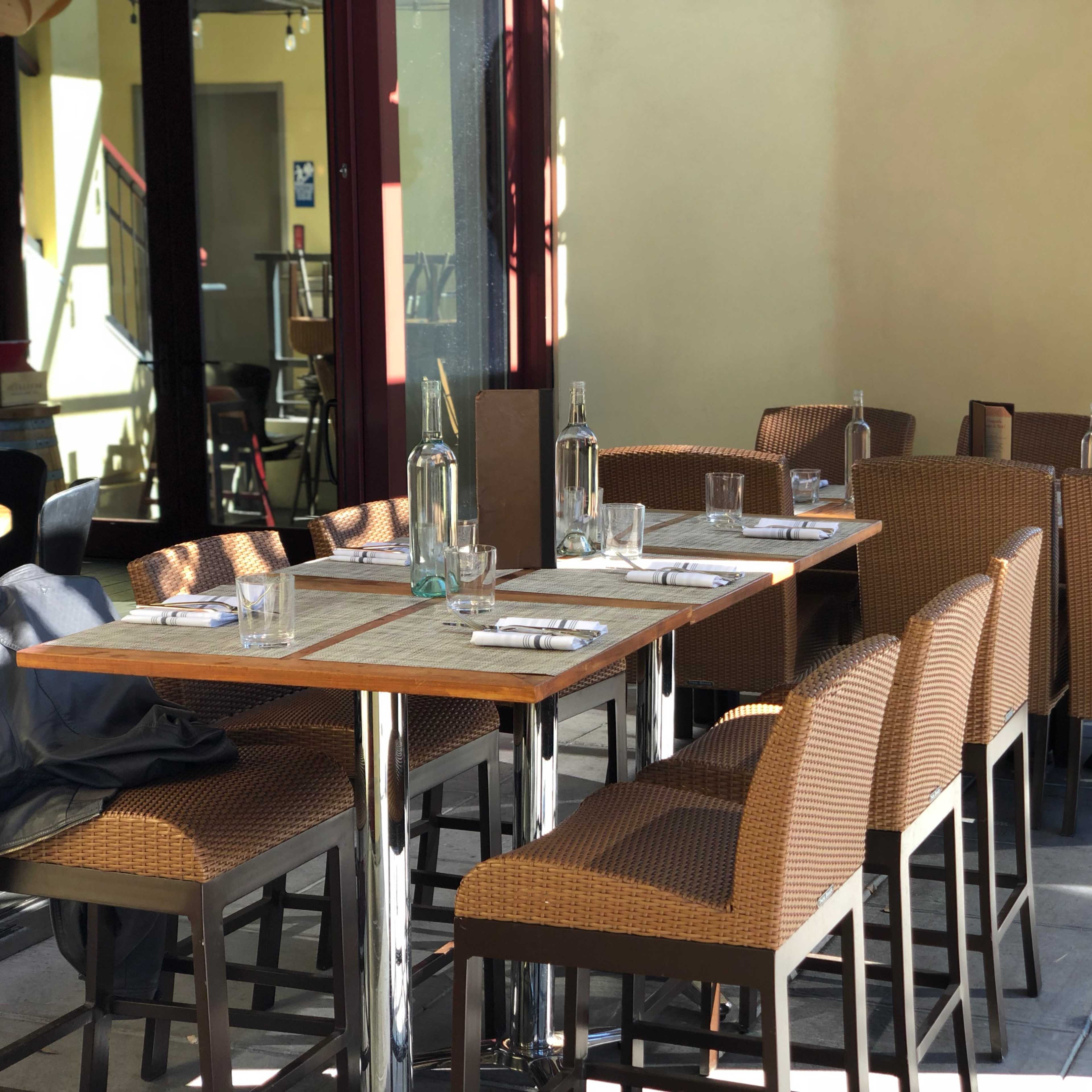 A dining patio features a set of empty, wicker-style chairs arranged around a large table, with glass bottles and napkins neatly placed on the table.