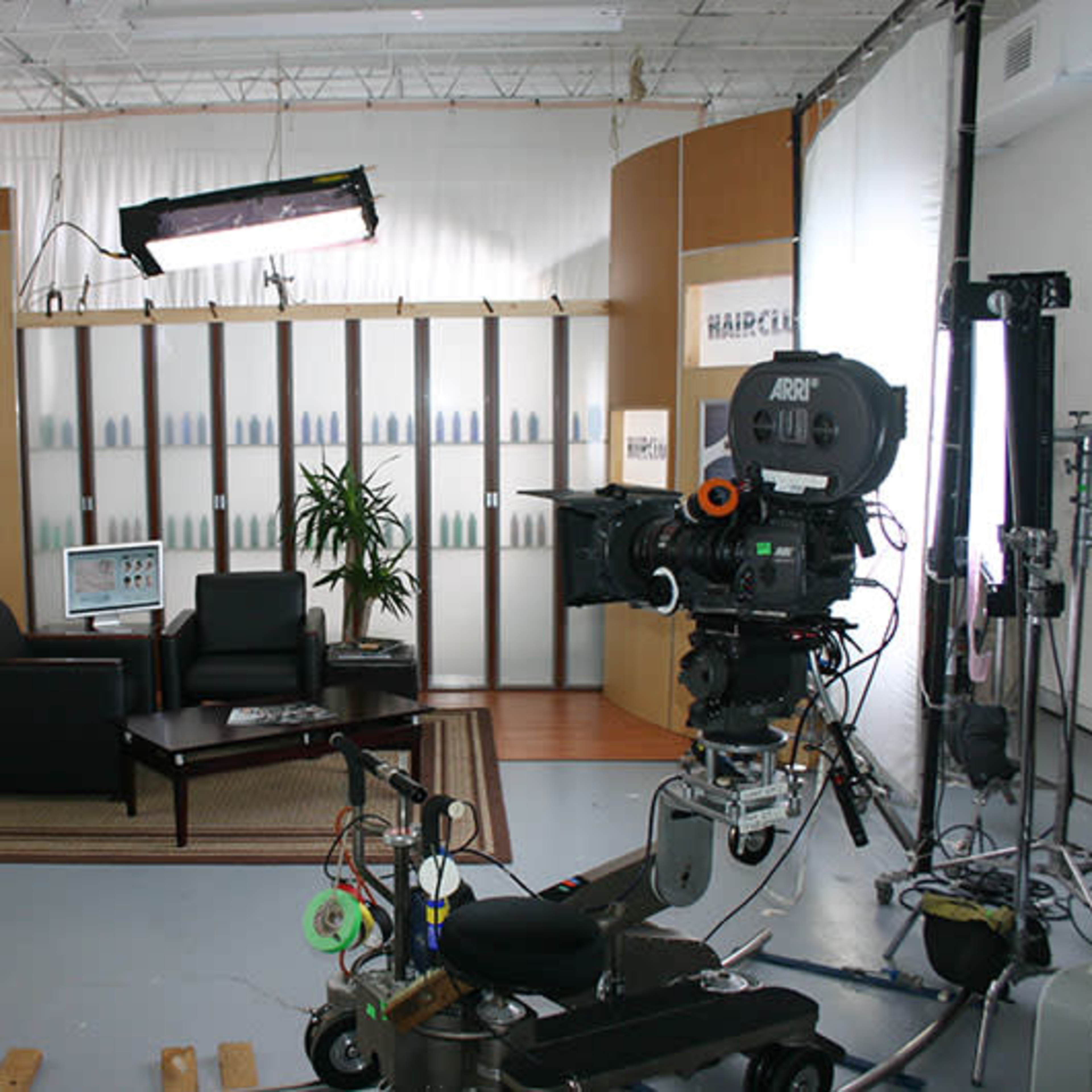 A television studio setup with a camera in the foreground, a seating area with two black chairs, and backdrop elements including a plant and a computer.