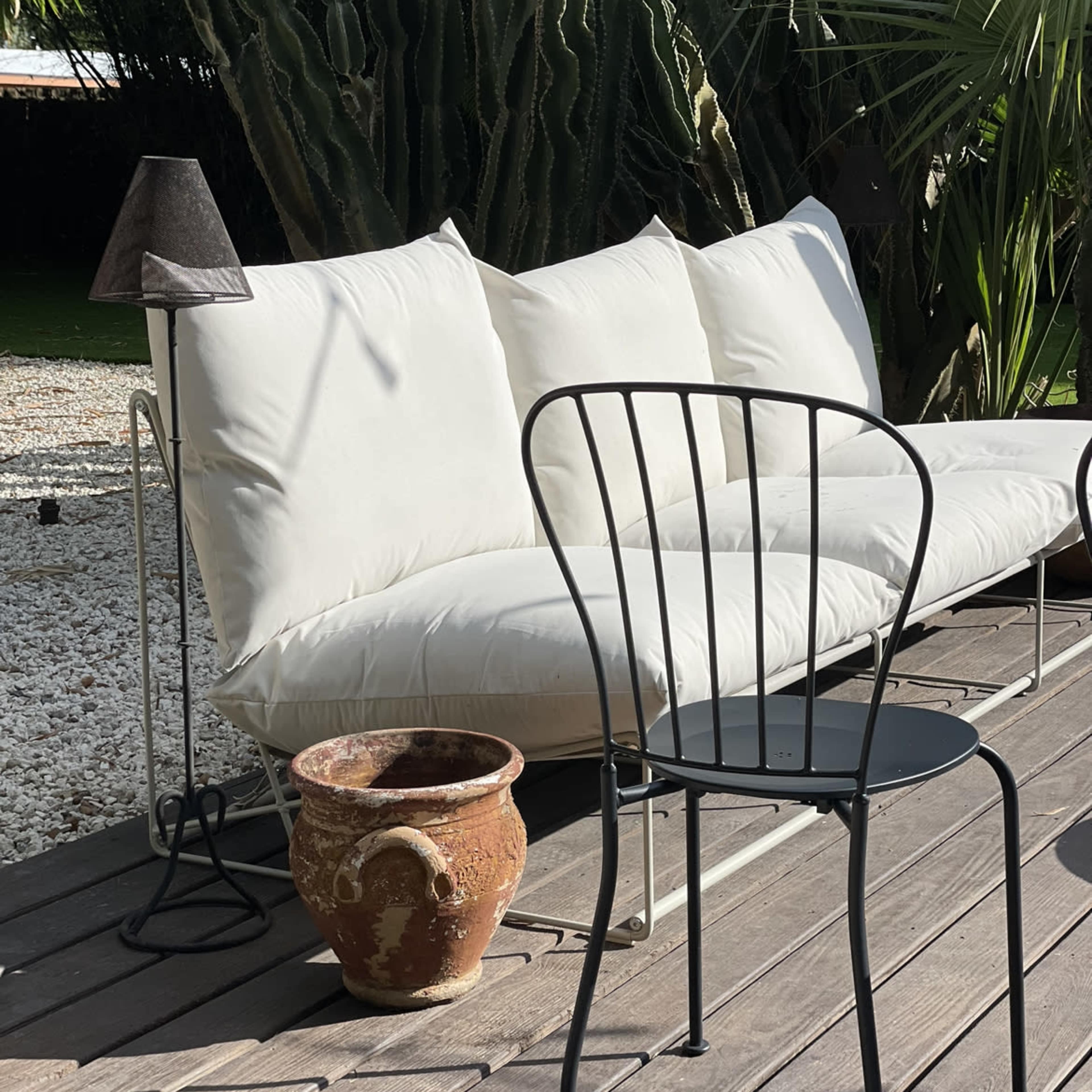 A white sofa and a black metal chair sit on a wooden deck surrounded by plants and a decorative pot.