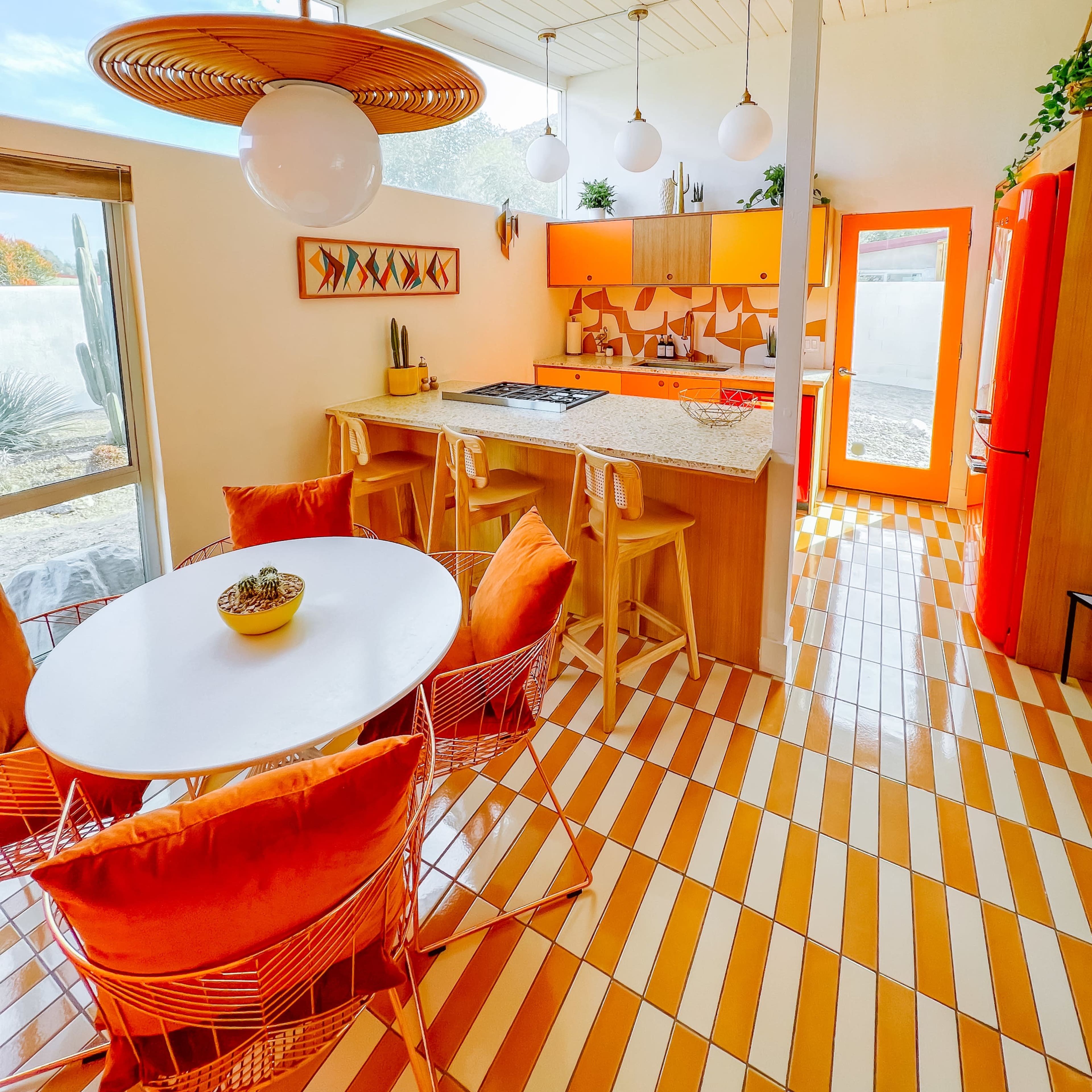 A brightly colored kitchen with orange cabinetry, a round white dining table surrounded by orange chairs, and a tiled floor in alternating stripes of orange and white.