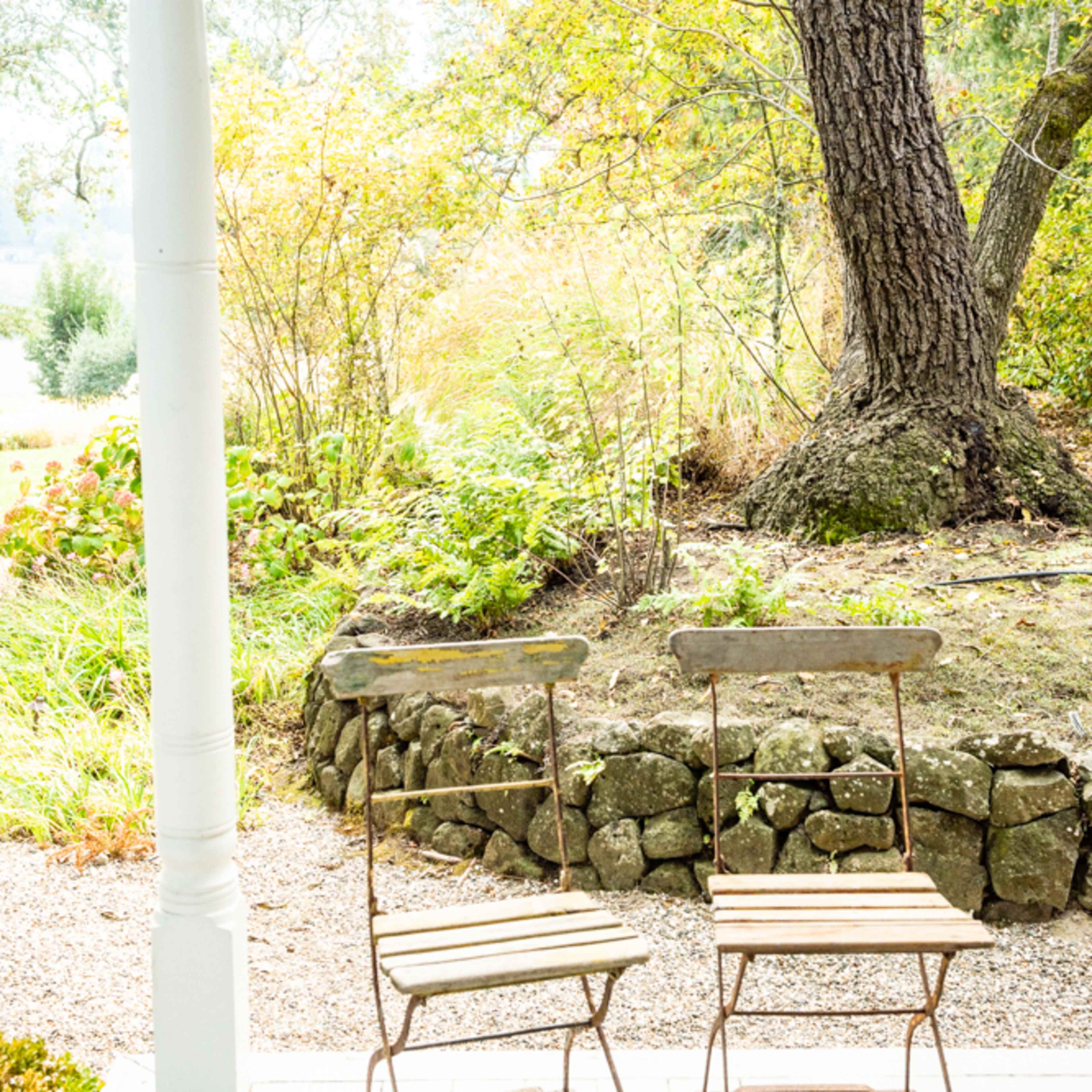 Two metal chairs stand on a porch, positioned near a stone wall and surrounded by greenery and a large tree.