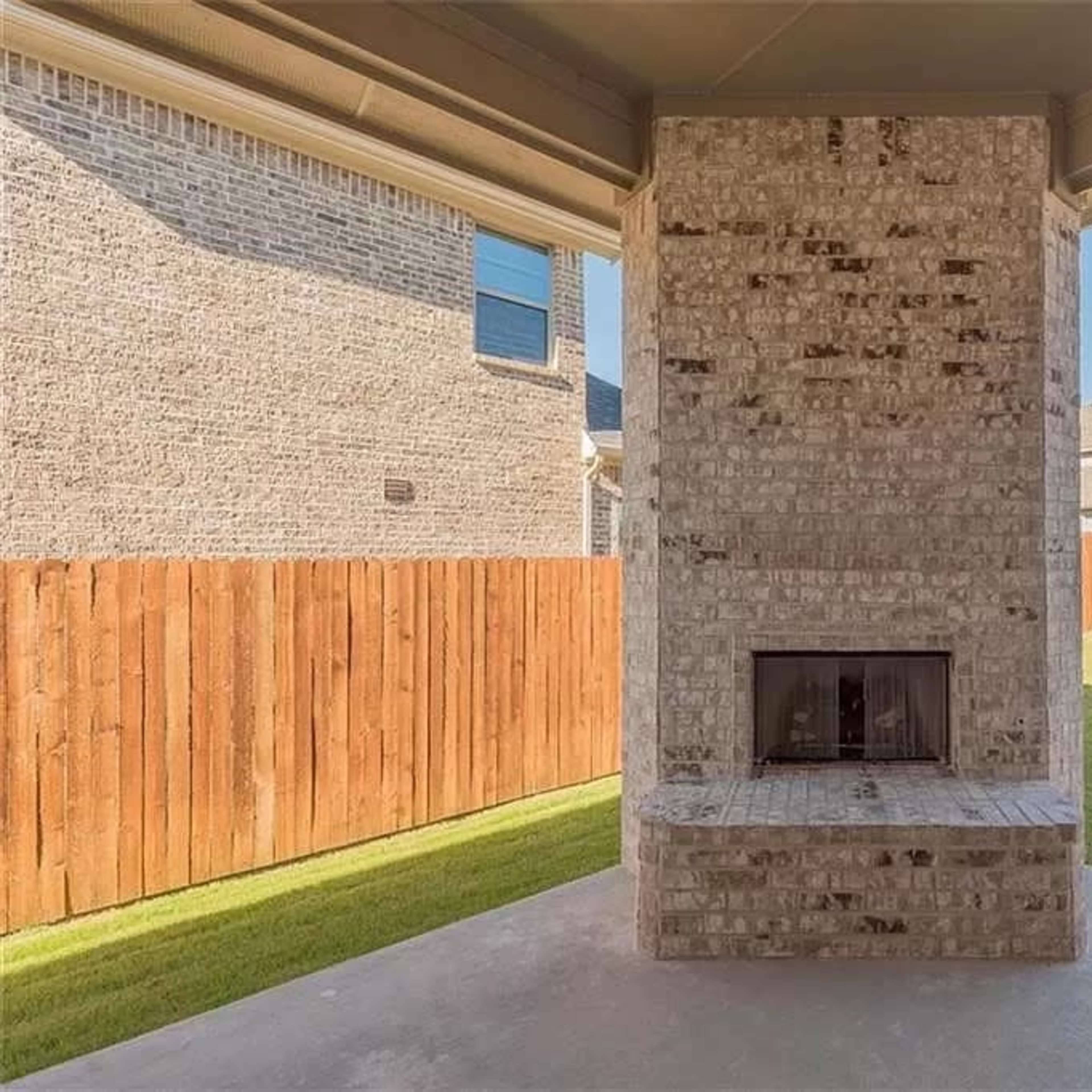 A brick outdoor fireplace situated on a concrete patio, surrounded by a grassy yard and a wooden fence.