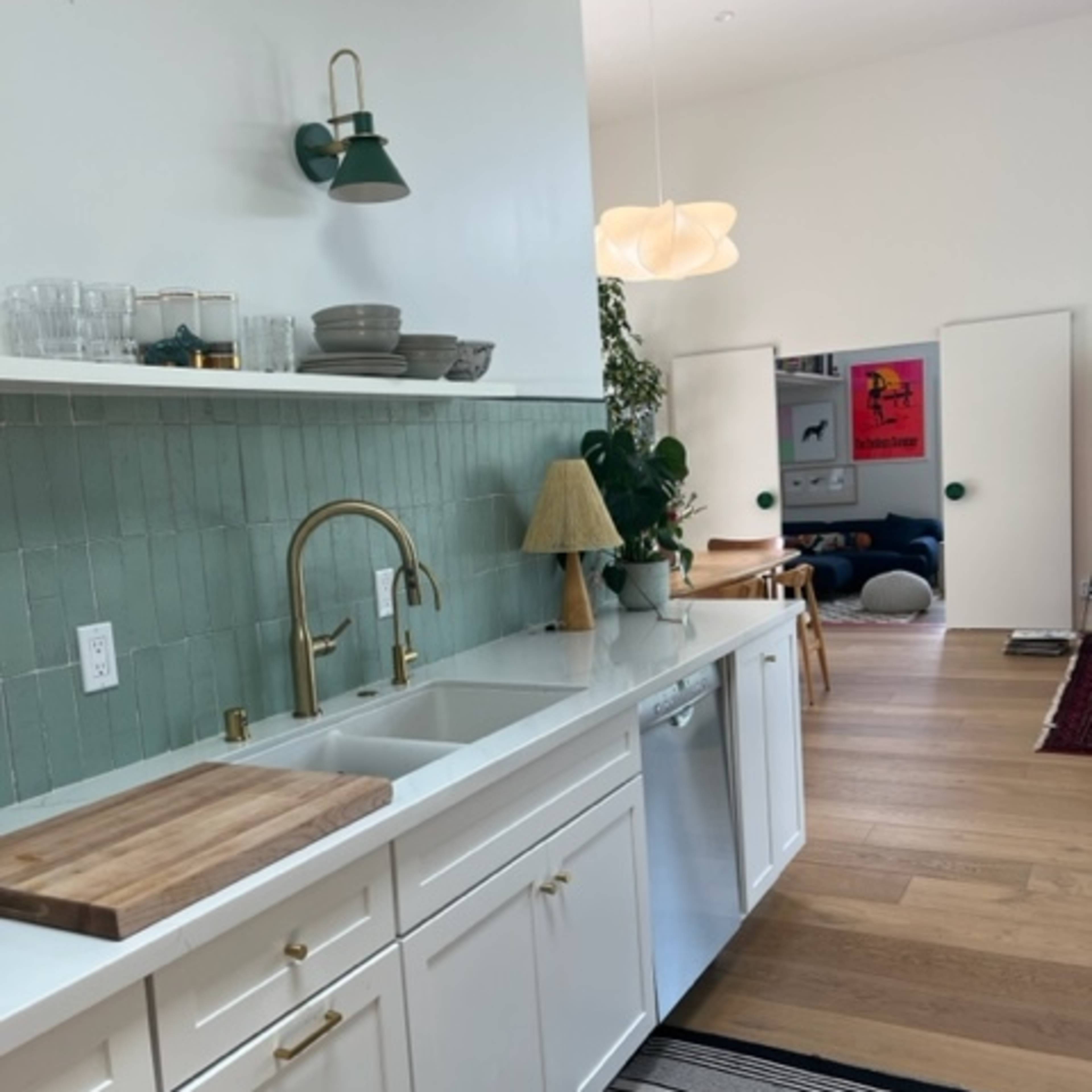 A modern kitchen with white cabinetry, a green tiled backsplash, a wooden cutting board on the counter, and a view into a living area featuring a colorful poster on the wall.