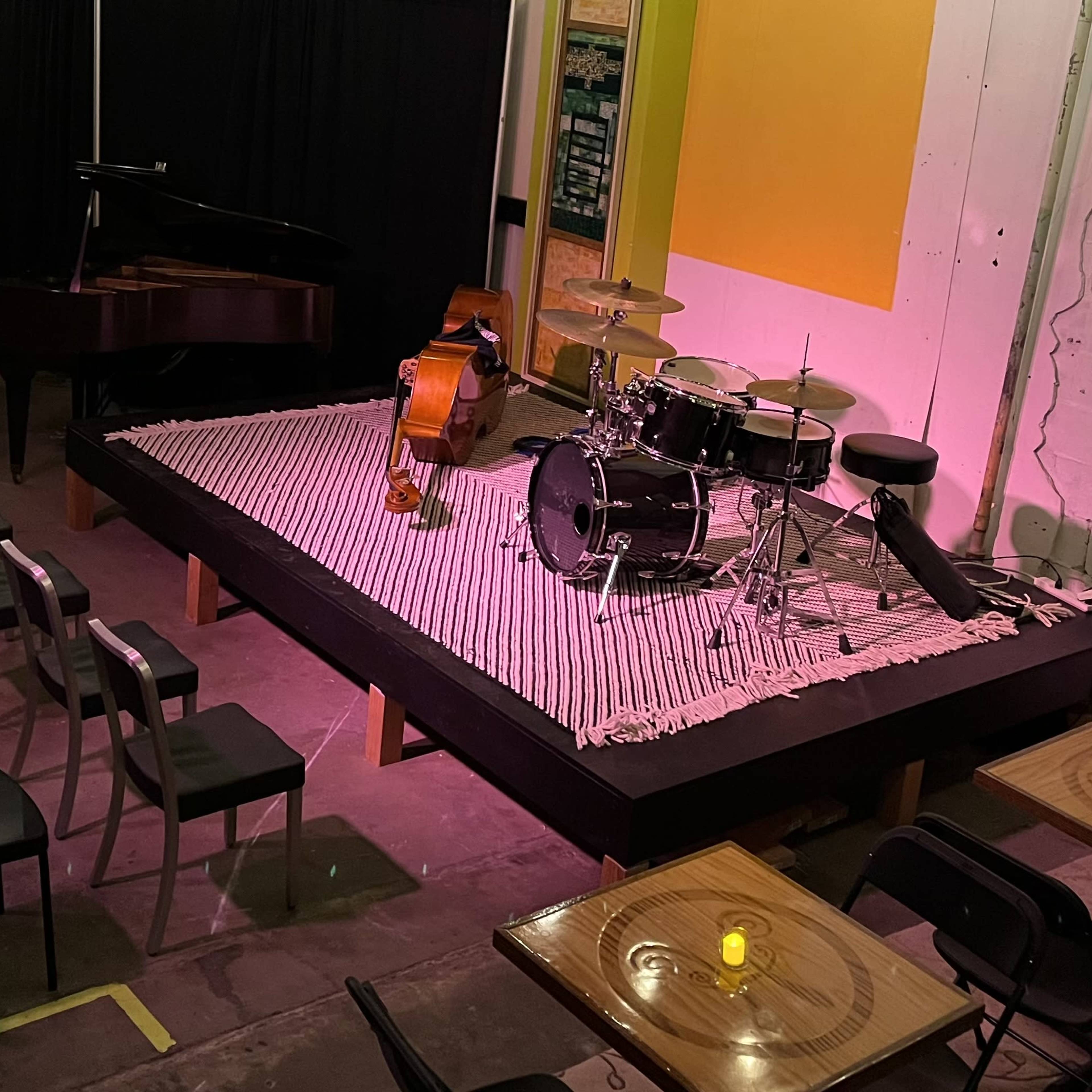 A small performance stage with a grand piano, double bass, drum set, and several rows of empty chairs arranged around tables.