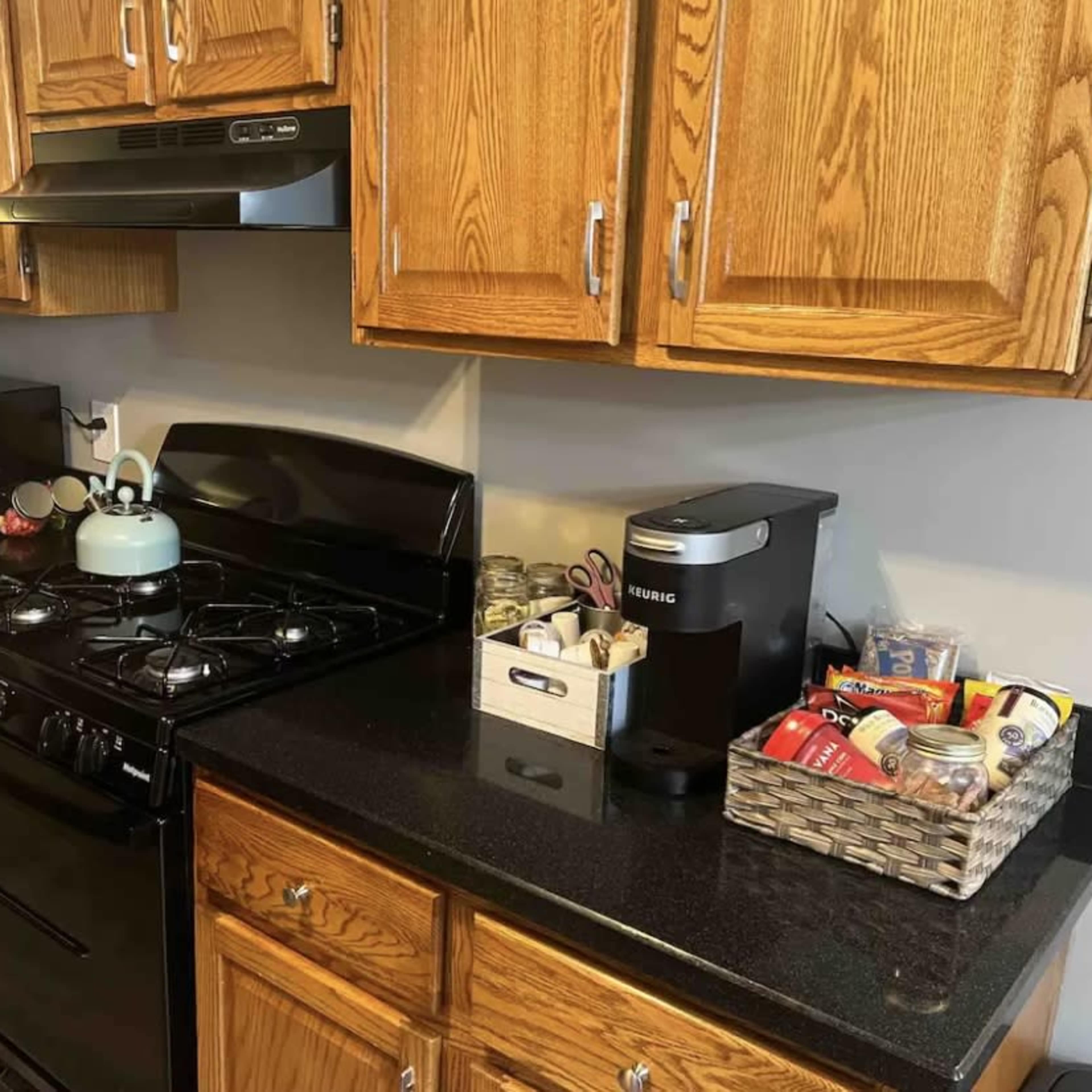 A kitchen counter with a black gas stove, a blue kettle, a microwave, and a coffee maker, surrounded by wooden cabinets and a basket containing various food items.