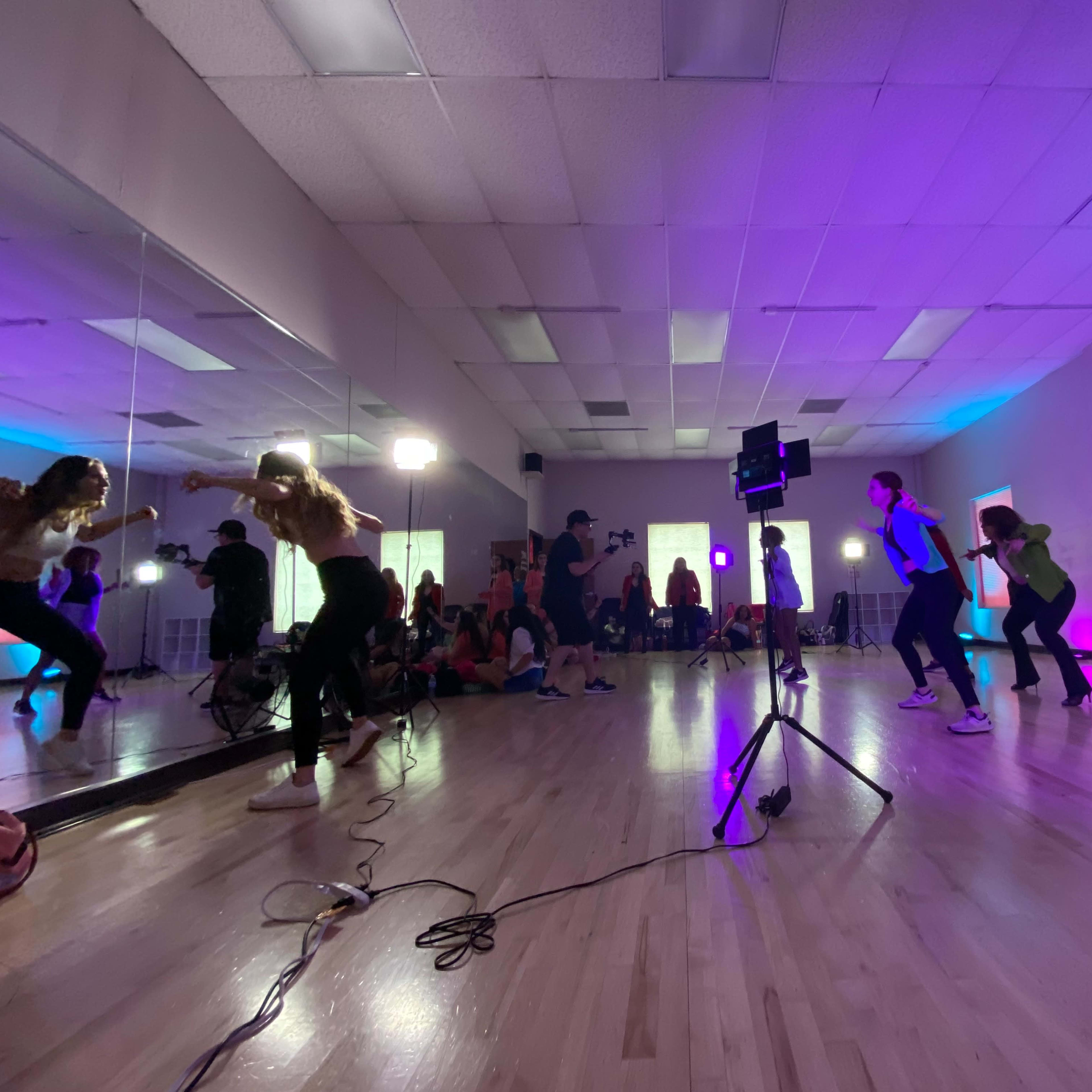 A group of people are dancing in a studio with mirrors, under colorful lights and photography equipment.