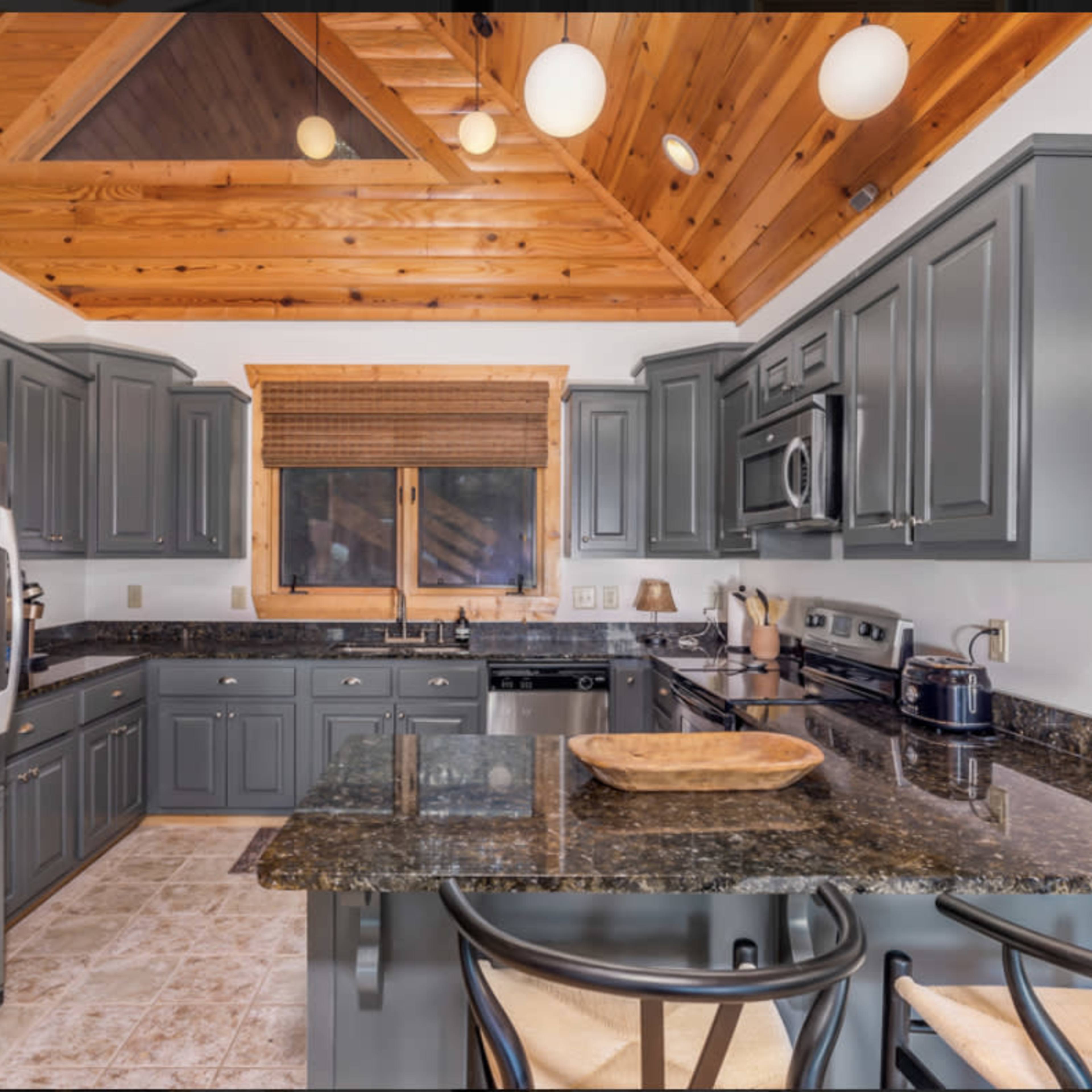 The kitchen features gray cabinets, a dark granite countertop, and wooden beams on the ceiling, with modern appliances and a large window.