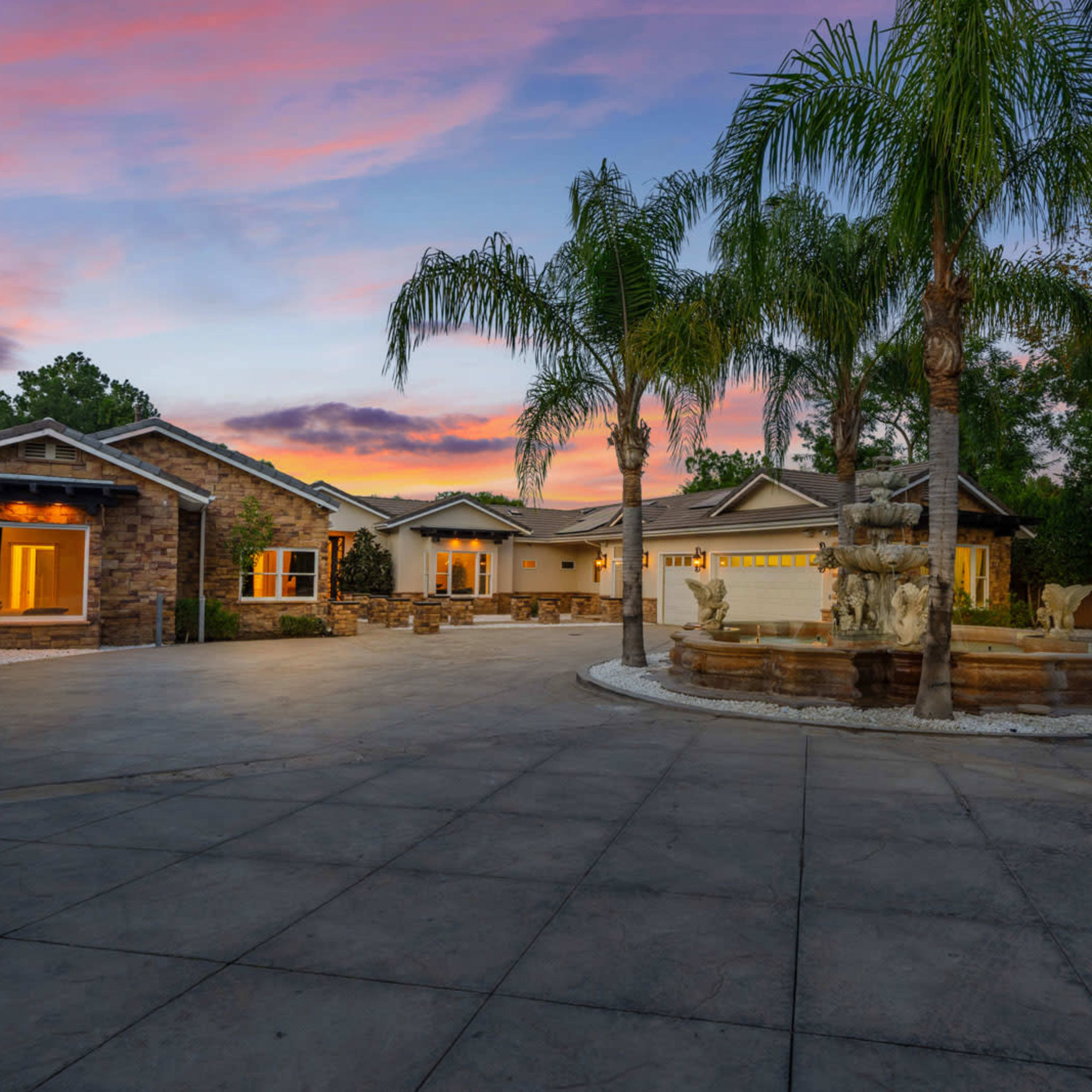 A modern home with a stone facade and landscaped front yard features a circular driveway and a fountain surrounded by palm trees at sunset.