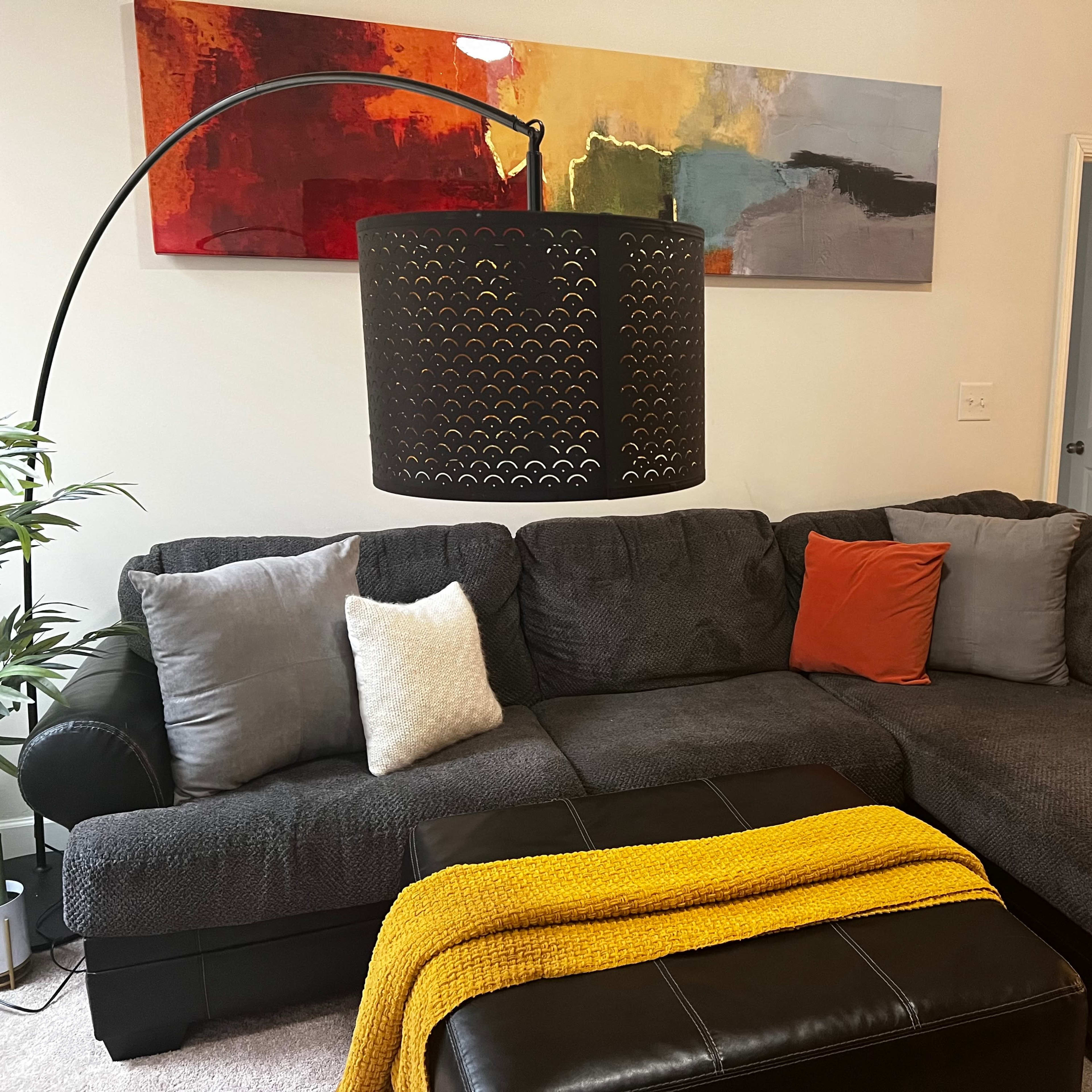 A gray sectional sofa with colorful throw pillows and a yellow blanket is positioned in a living room under a black floor lamp, with abstract artwork on the wall.
