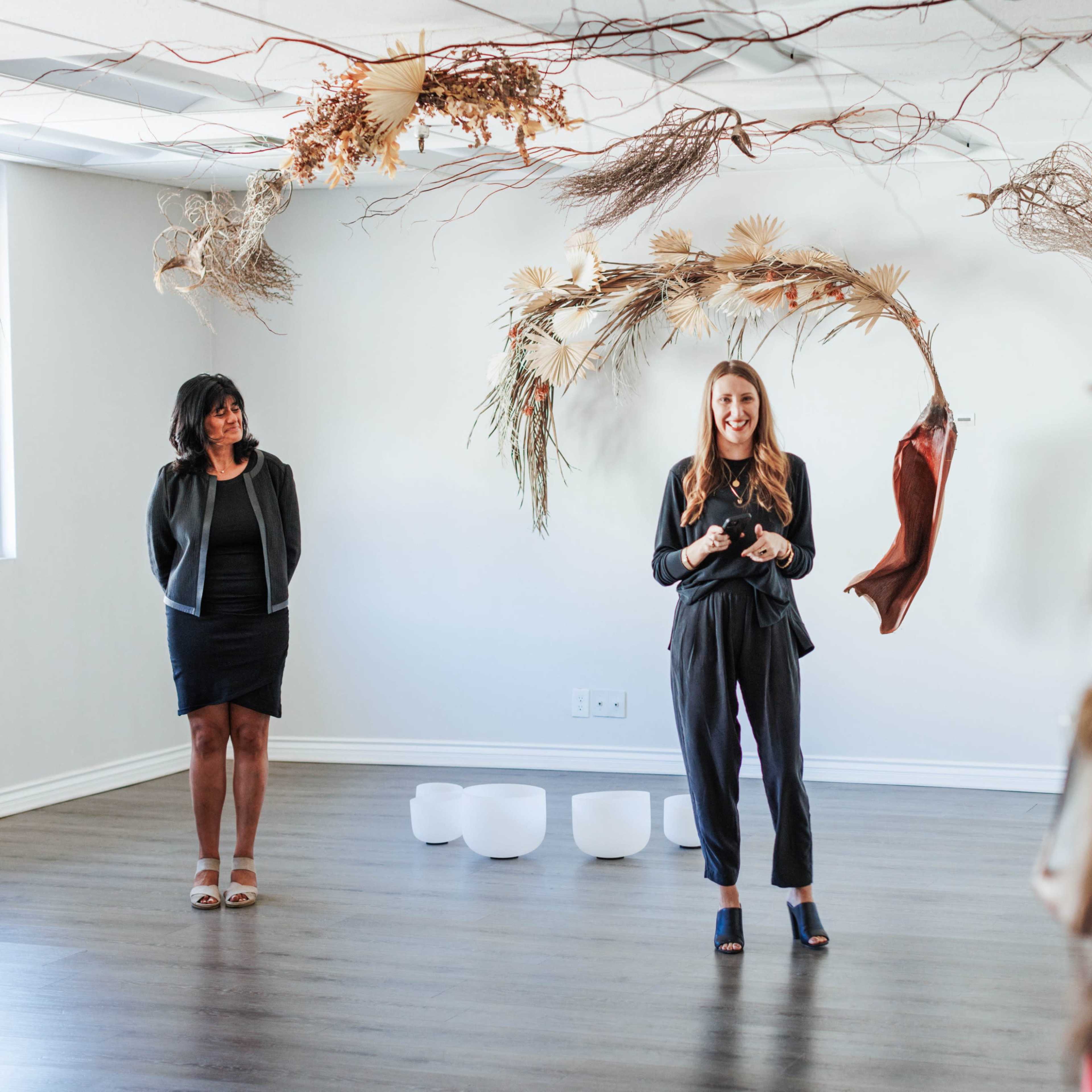 A woman in a black outfit stands speaking in front of a small audience, while another woman stands to her left, with decorative branches and materials hanging from the ceiling.