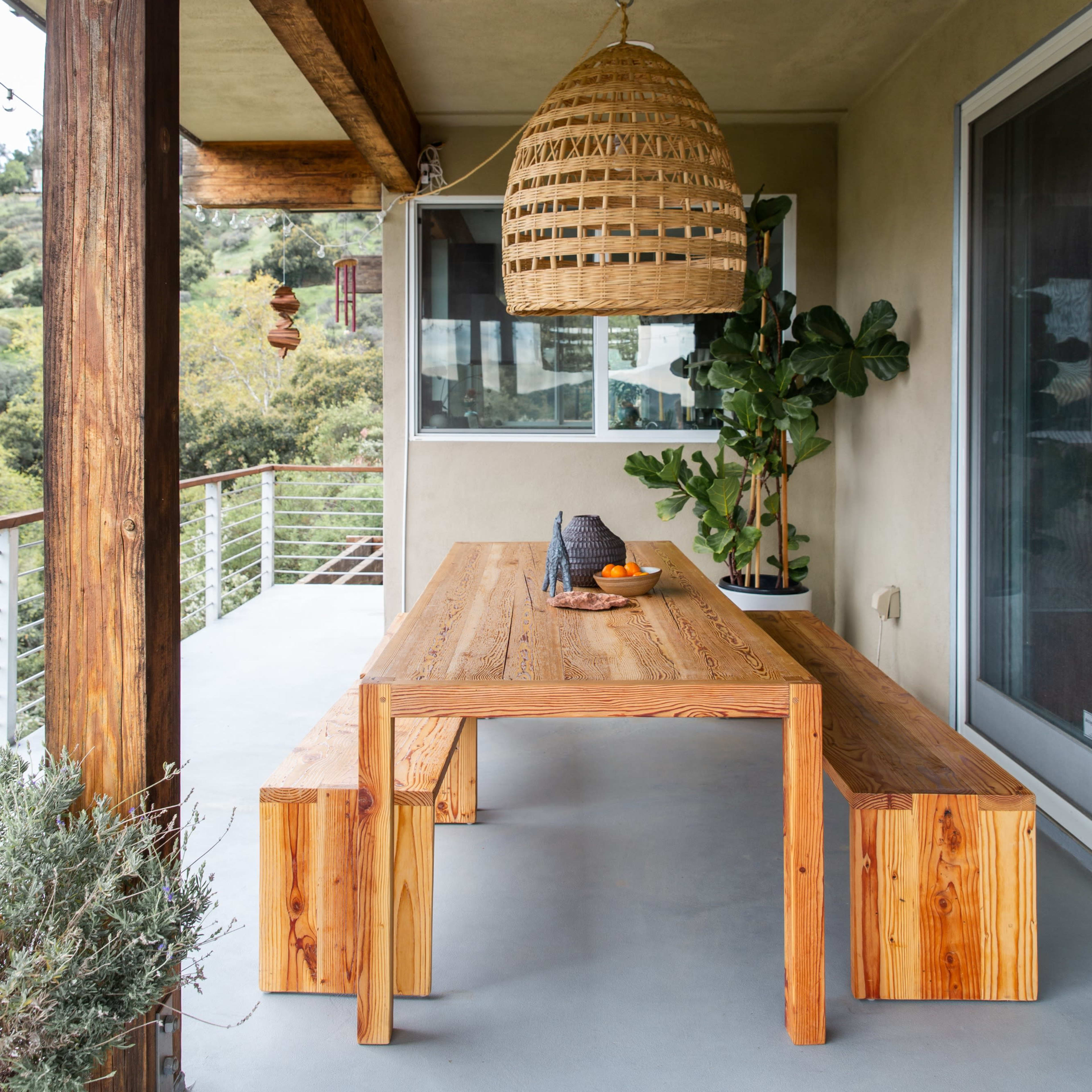 A wooden table with benches sits on a patio under a woven pendant light, surrounded by greenery and a building with large windows.