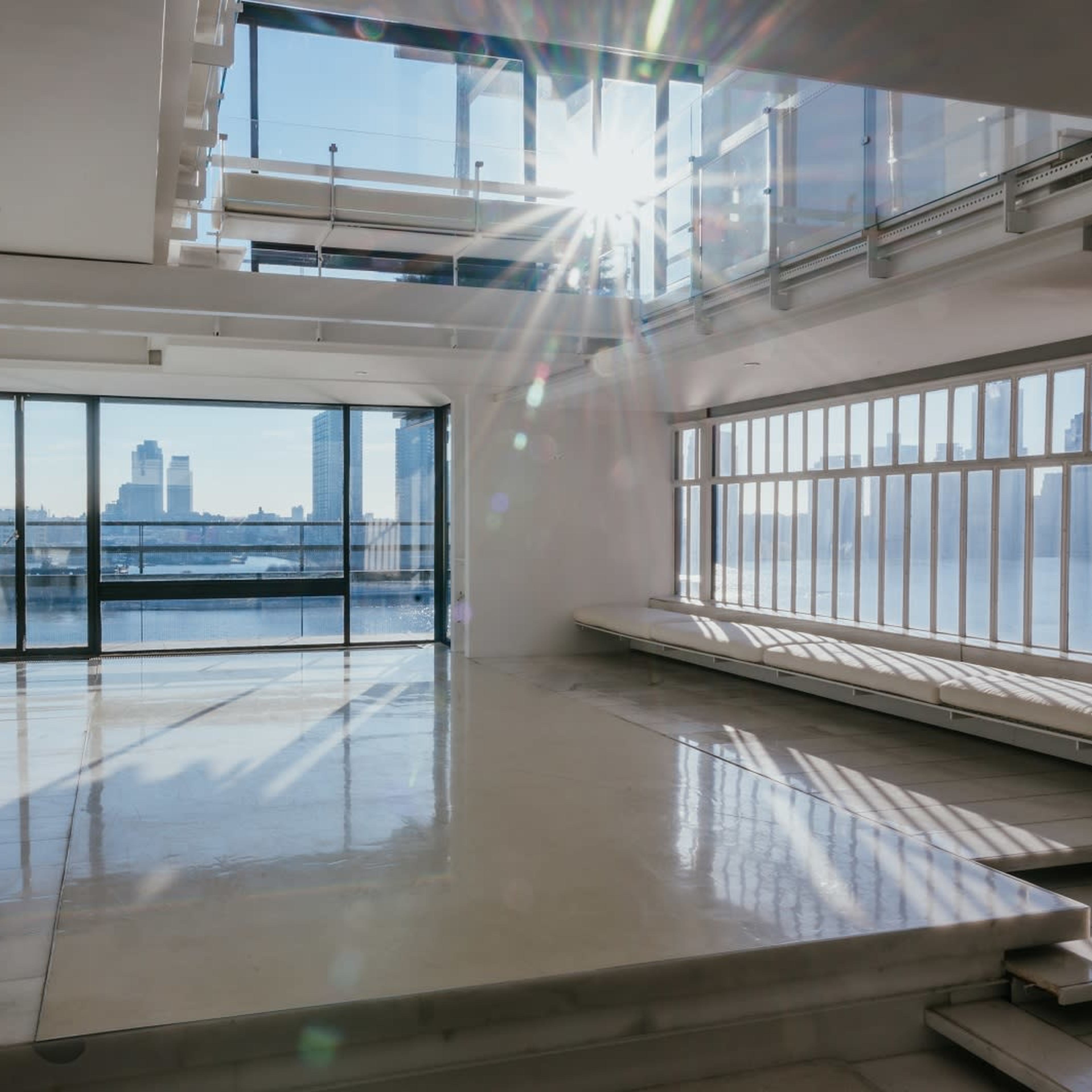 A spacious, modern interior with large windows allowing natural light to illuminate the room and a view of a river and skyline beyond.