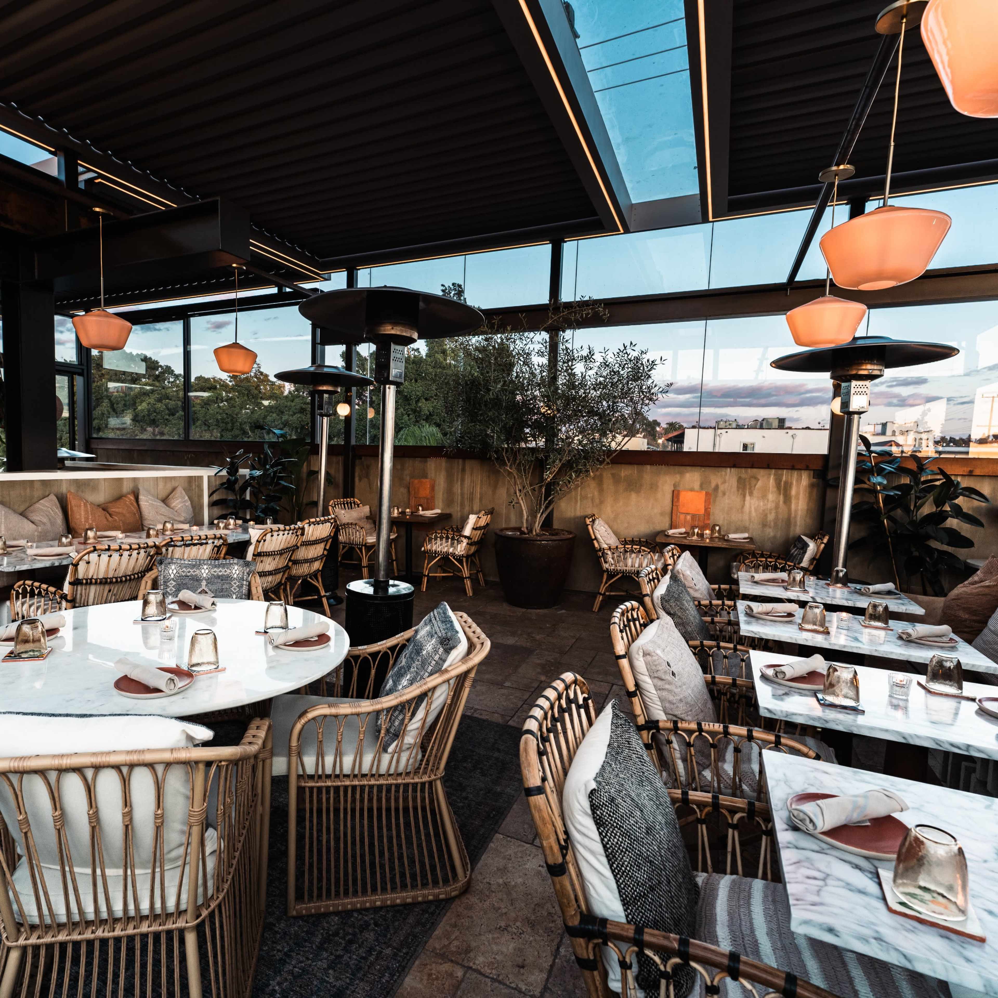 A modern restaurant with tables set for dining, featuring wicker chairs and large windows that provide ample natural light.