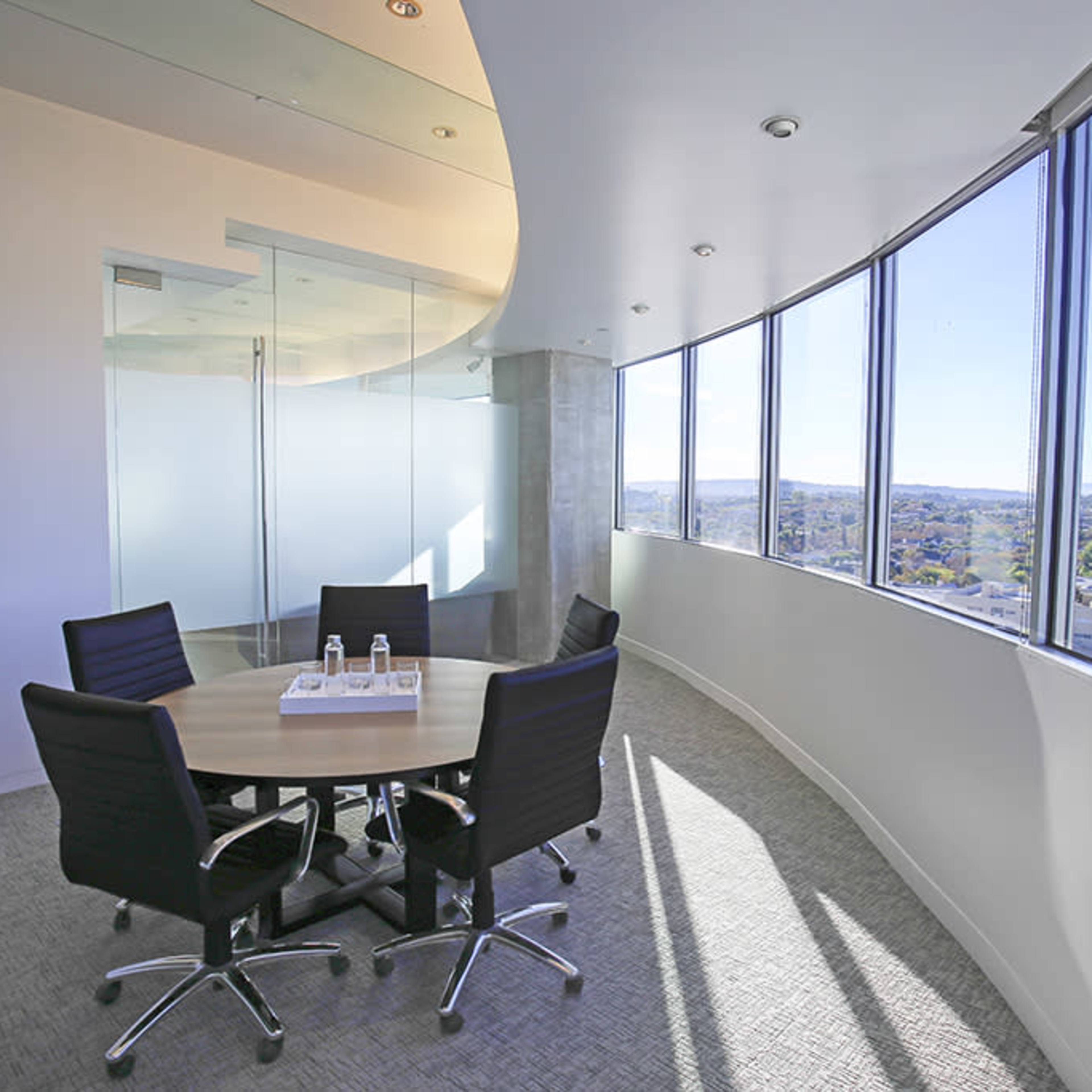 A modern conference room with a round table and four black chairs, featuring large windows that offer a view of the surrounding landscape.