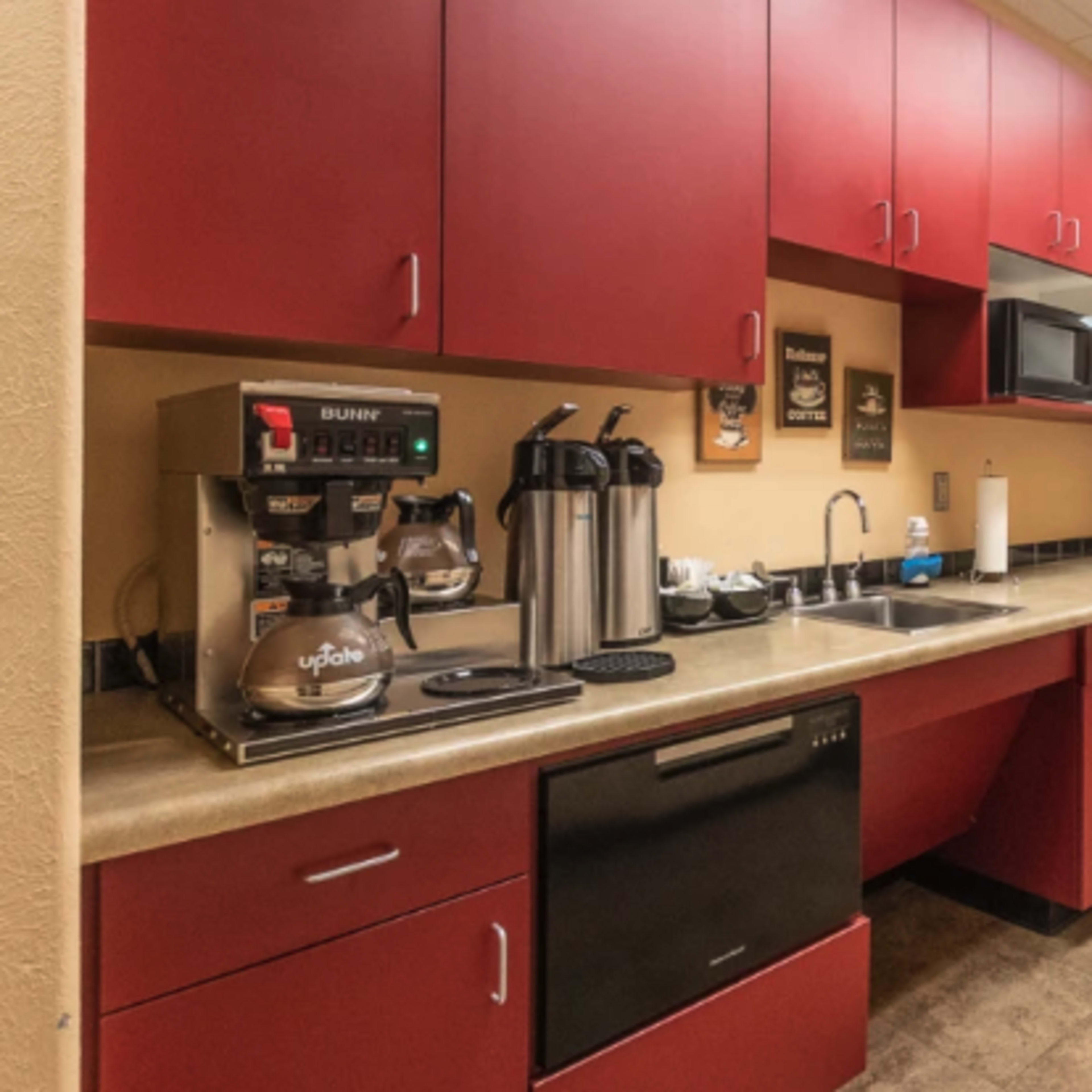 A kitchen area featuring red cabinets, a coffee machine, microwave, and a black refrigerator, adjacent to a hallway.