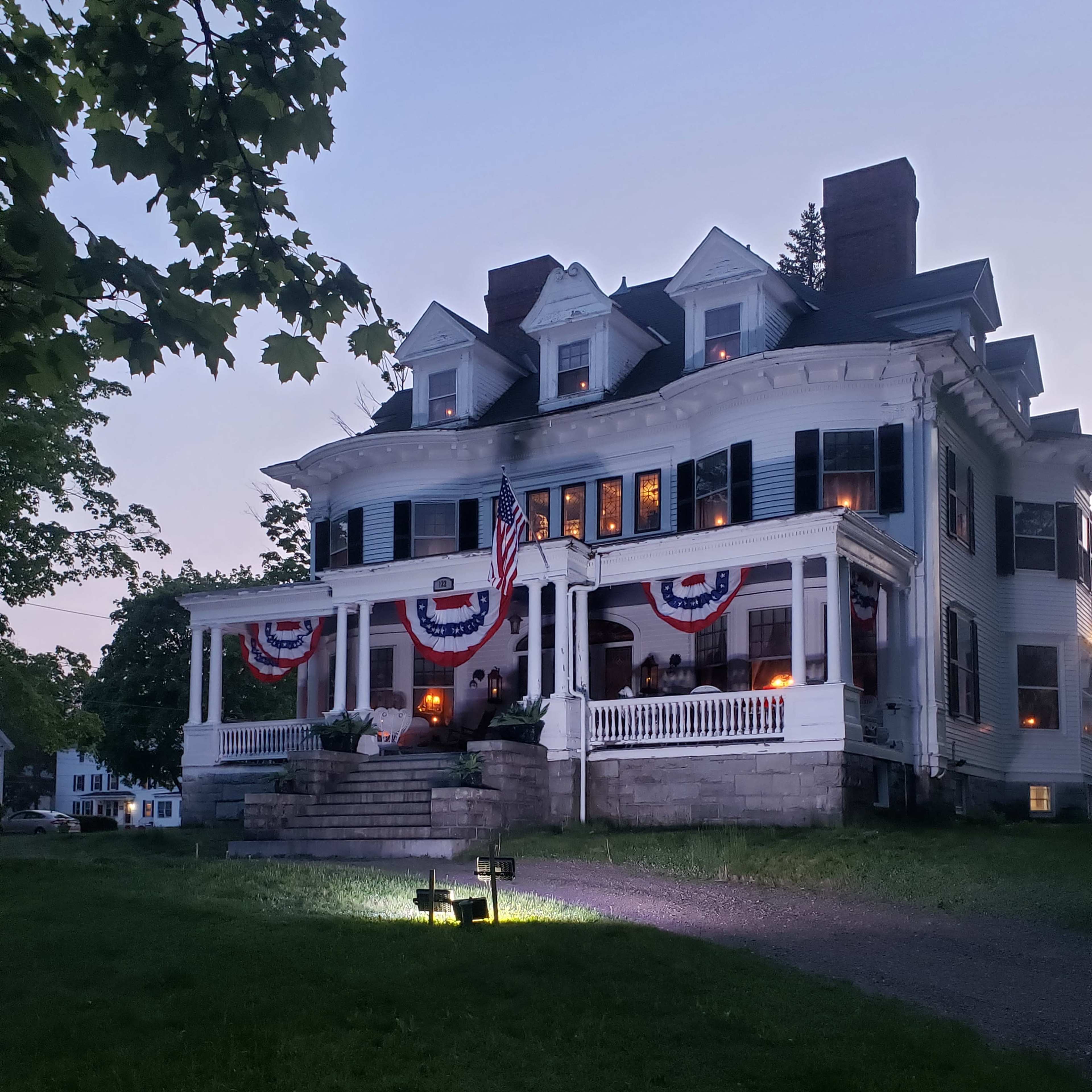 A large, historic house is adorned with patriotic bunting and illuminated by outdoor lighting during twilight.