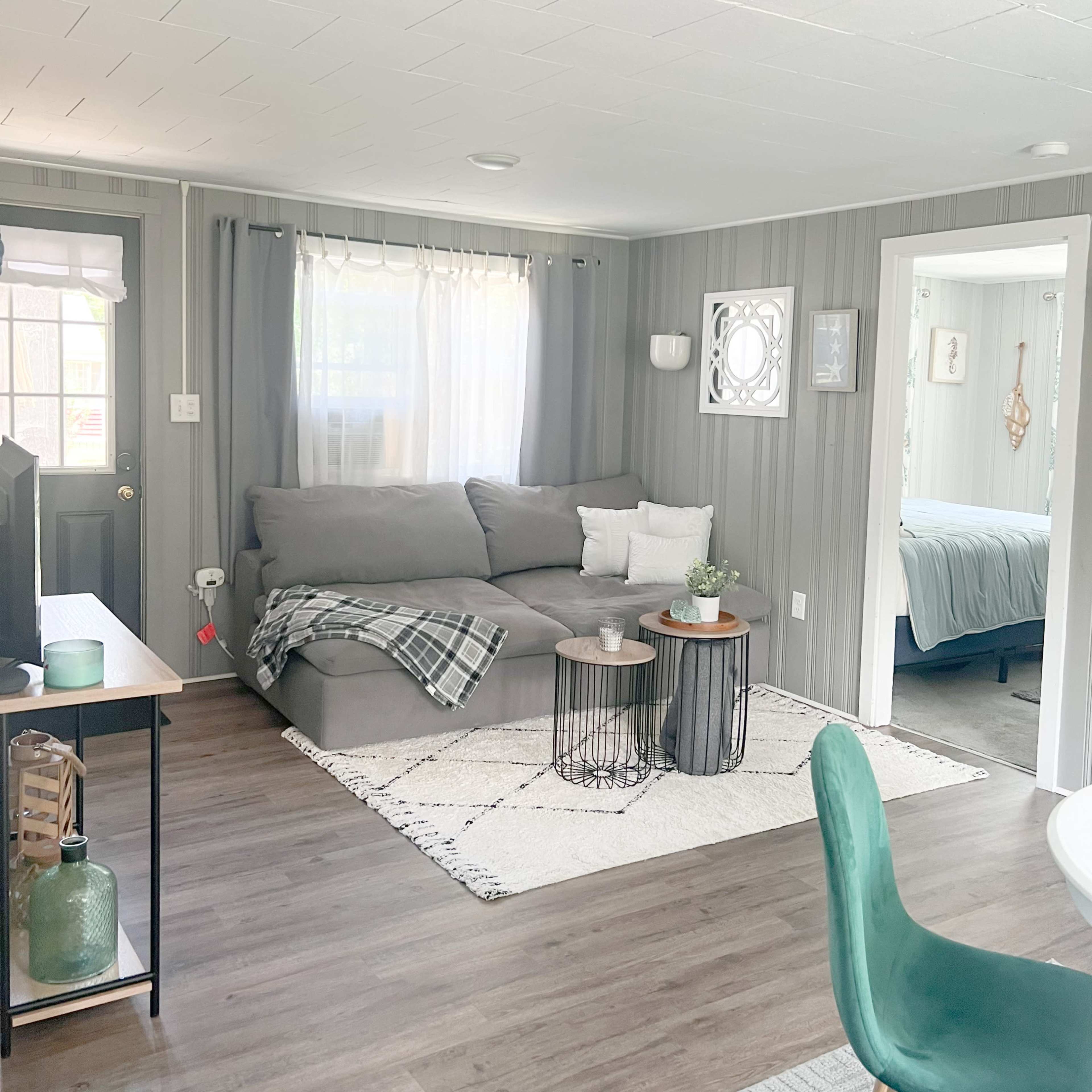A living room with a gray sectional couch, two side tables, a decorative rug, and a view into a bedroom in the background.