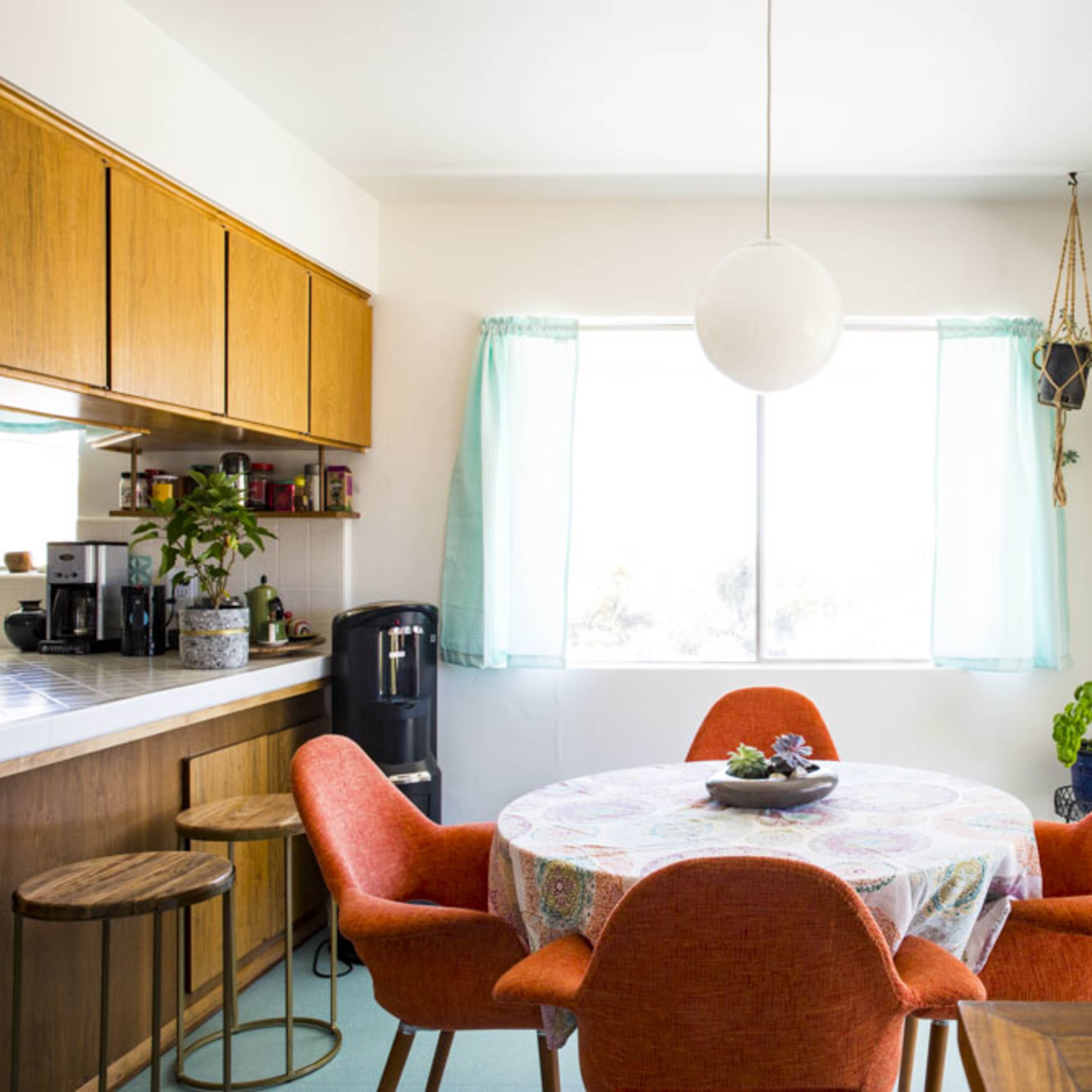 A dining area features a round table with a colorful tablecloth surrounded by four orange chairs, next to a window with sheer curtains and a sliding glass door.