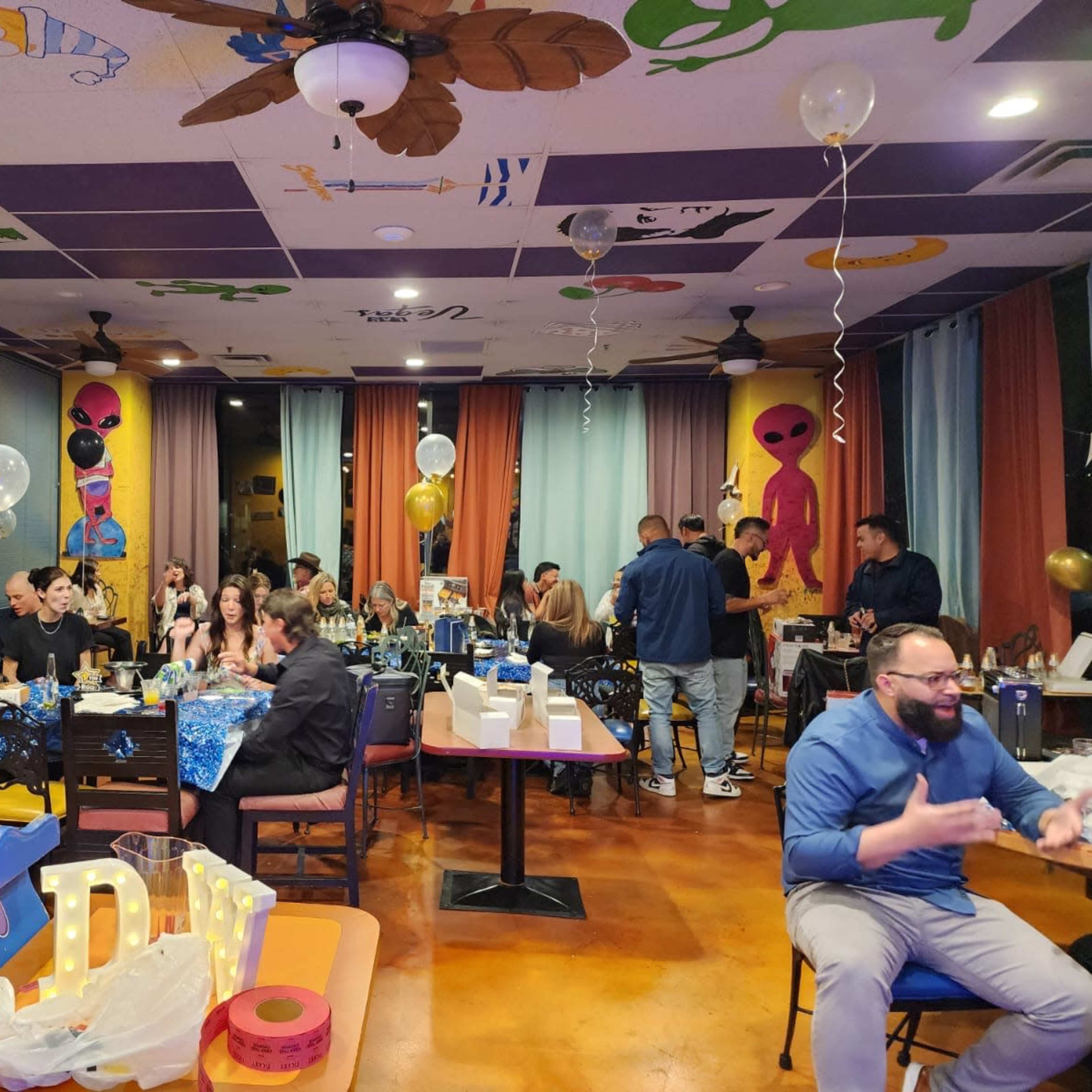A group of people celebrates an event in a decorated room filled with balloons and tables covered in party supplies.
