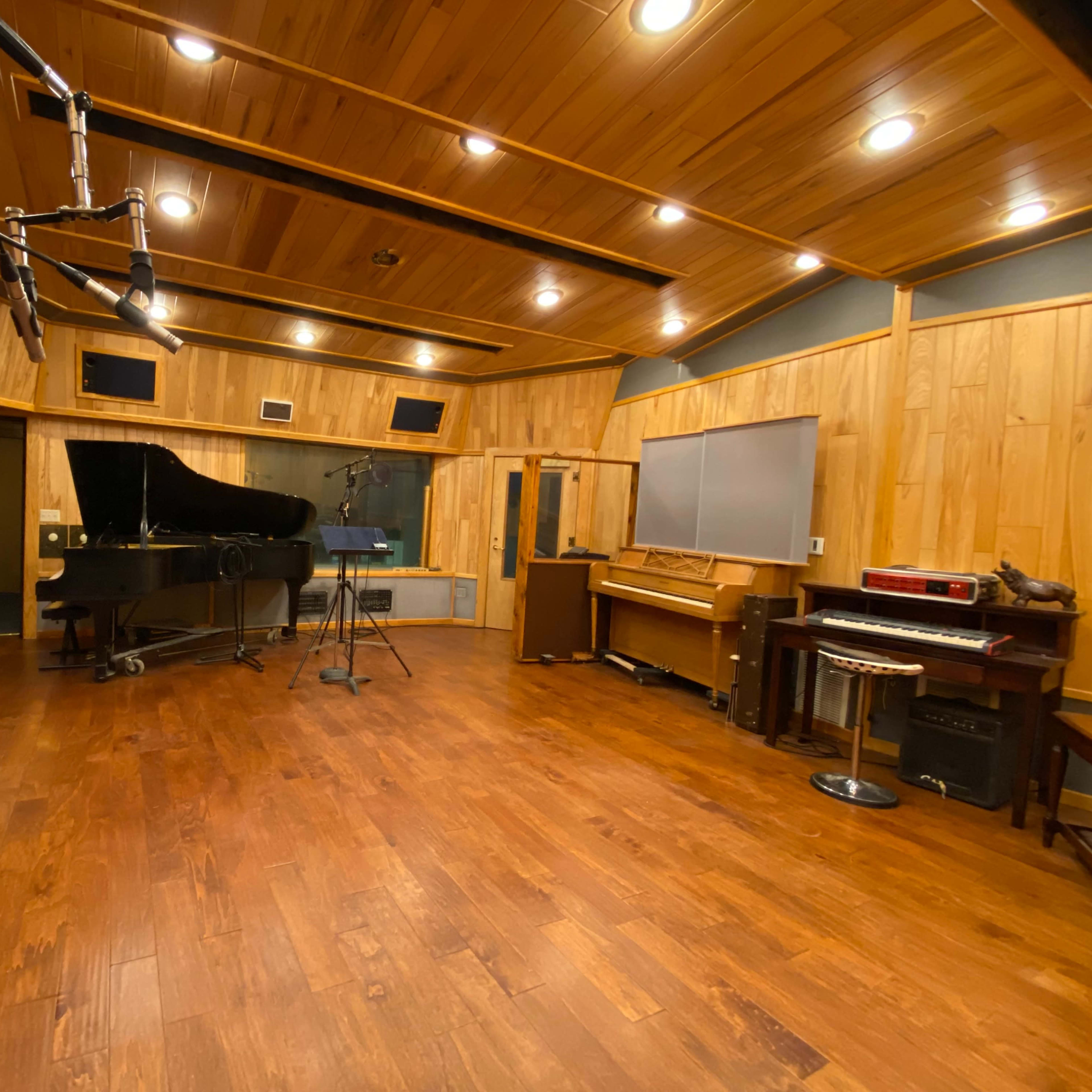 A music studio featuring a grand piano, an upright piano, and various recording equipment in a wooden-paneled room.