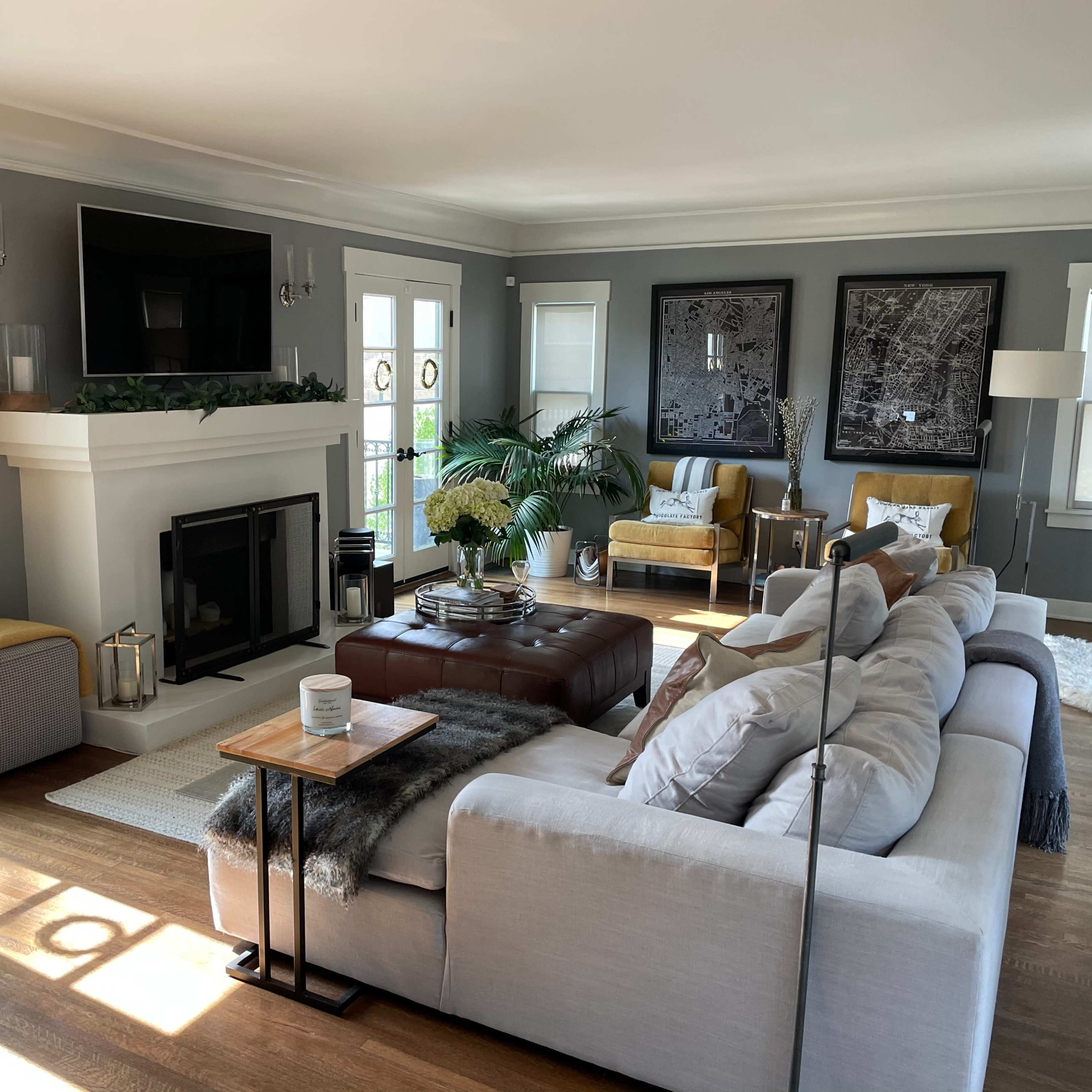 The living room features a neutral color palette with a large sectional sofa, a coffee table, a fireplace, and two armchairs, all arranged around a central area with natural light coming in through the windows.