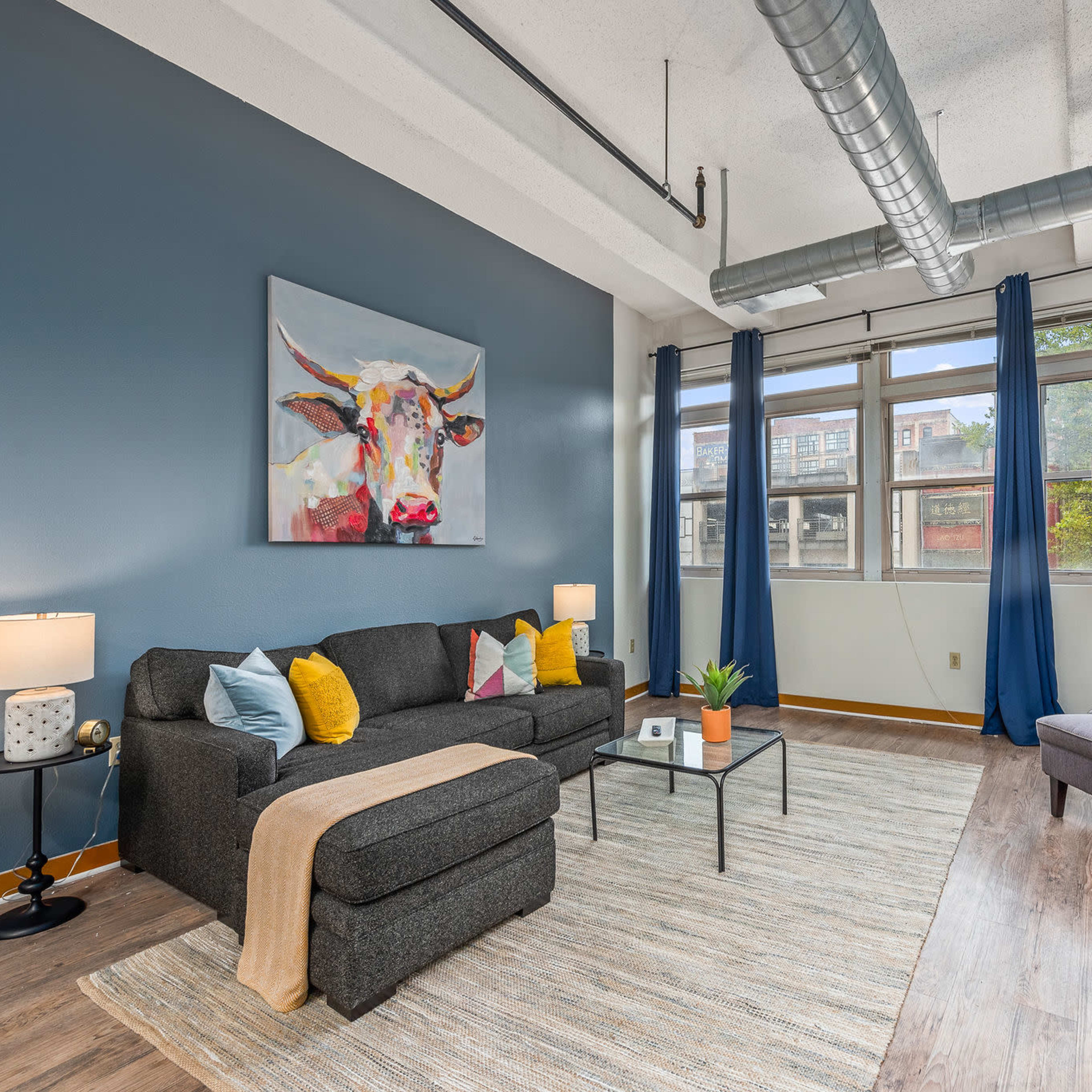 A modern living room with a gray sofa, yellow and blue accent pillows, a coffee table, and a large window with blue curtains, featuring a painting of a cow on the wall.