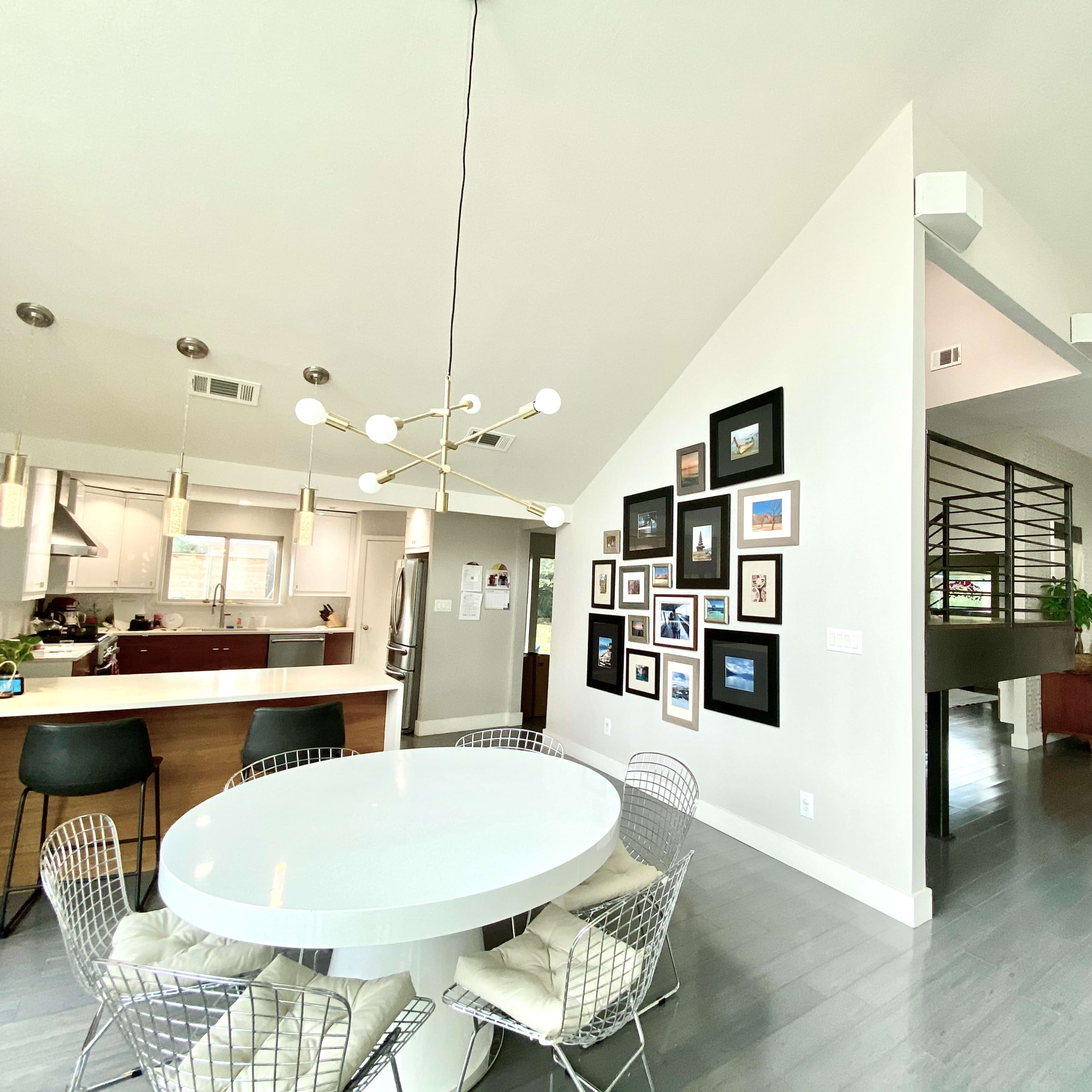 A spacious, modern home interior features a round white dining table with wire chairs, a gallery wall of framed pictures, and a large open kitchen visible in the background.