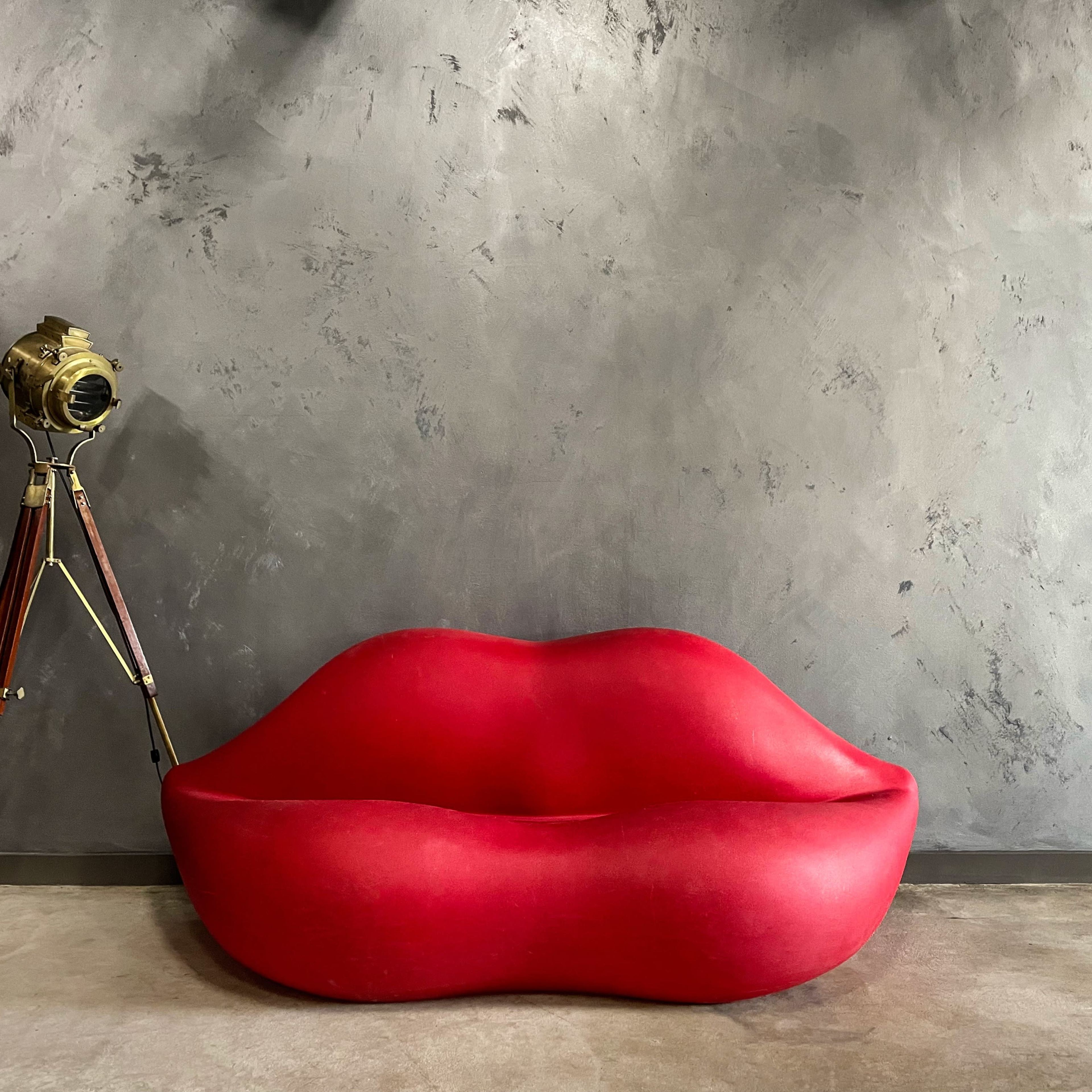 A red, lip-shaped sofa is placed against a textured gray wall next to a vintage-style tripod camera.
