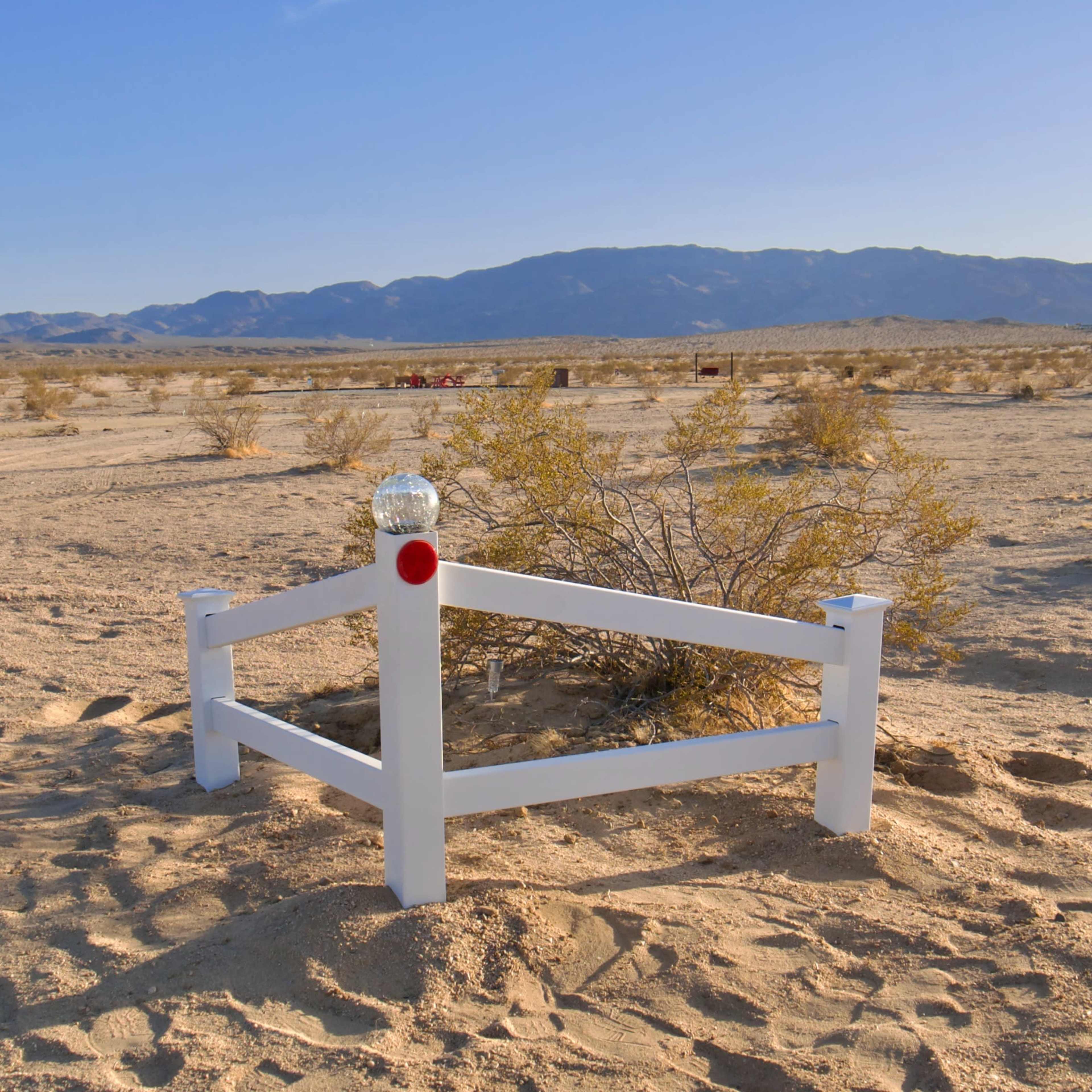 A white wooden fence surrounds a small post with a reflective glass orb, set against a desolate desert landscape with distant mountains.
