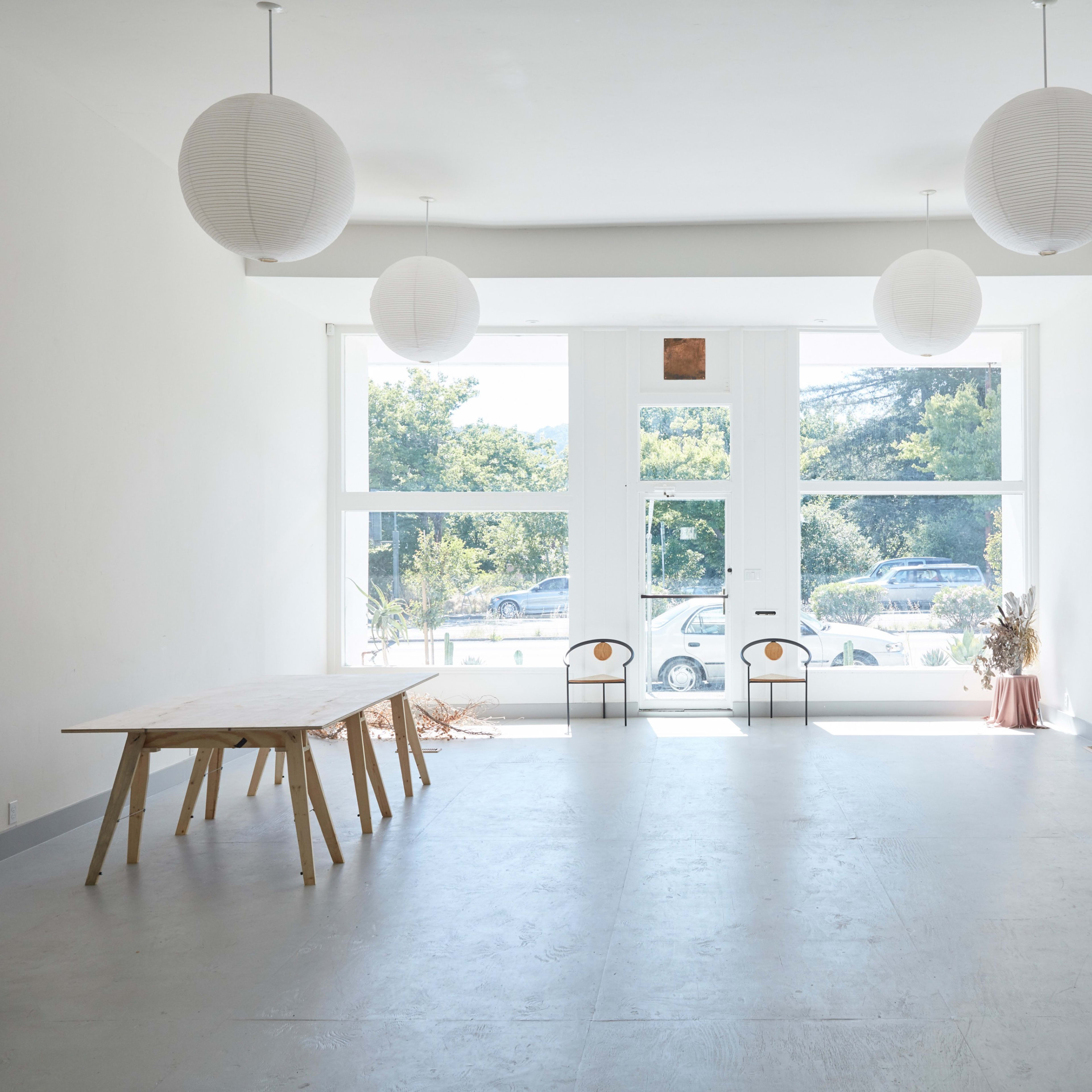 A spacious, minimalist room with large windows, a wooden table, and three paper lanterns hanging from the ceiling.