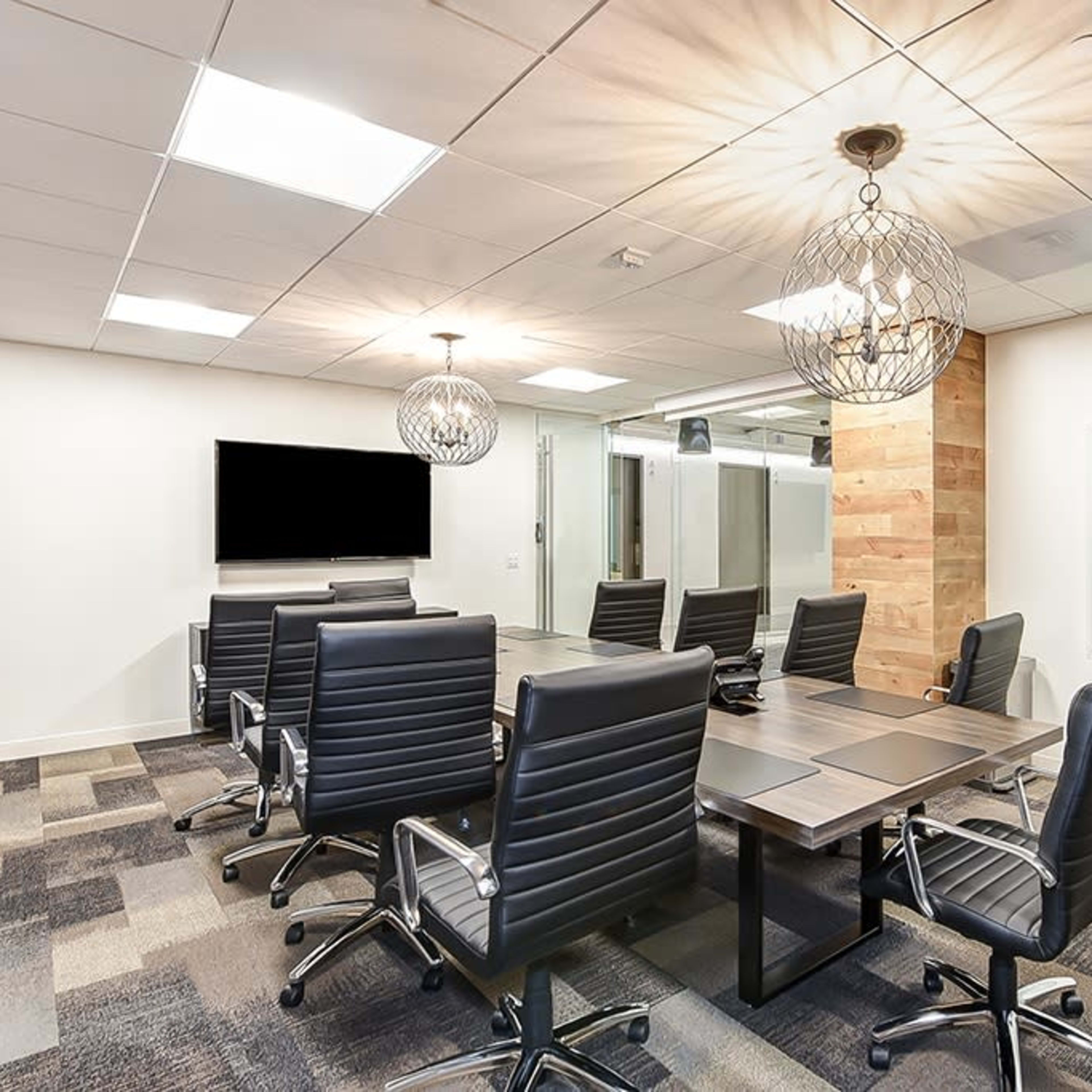 A modern conference room features a large table surrounded by leather chairs, with a wall-mounted screen and decorative light fixtures.