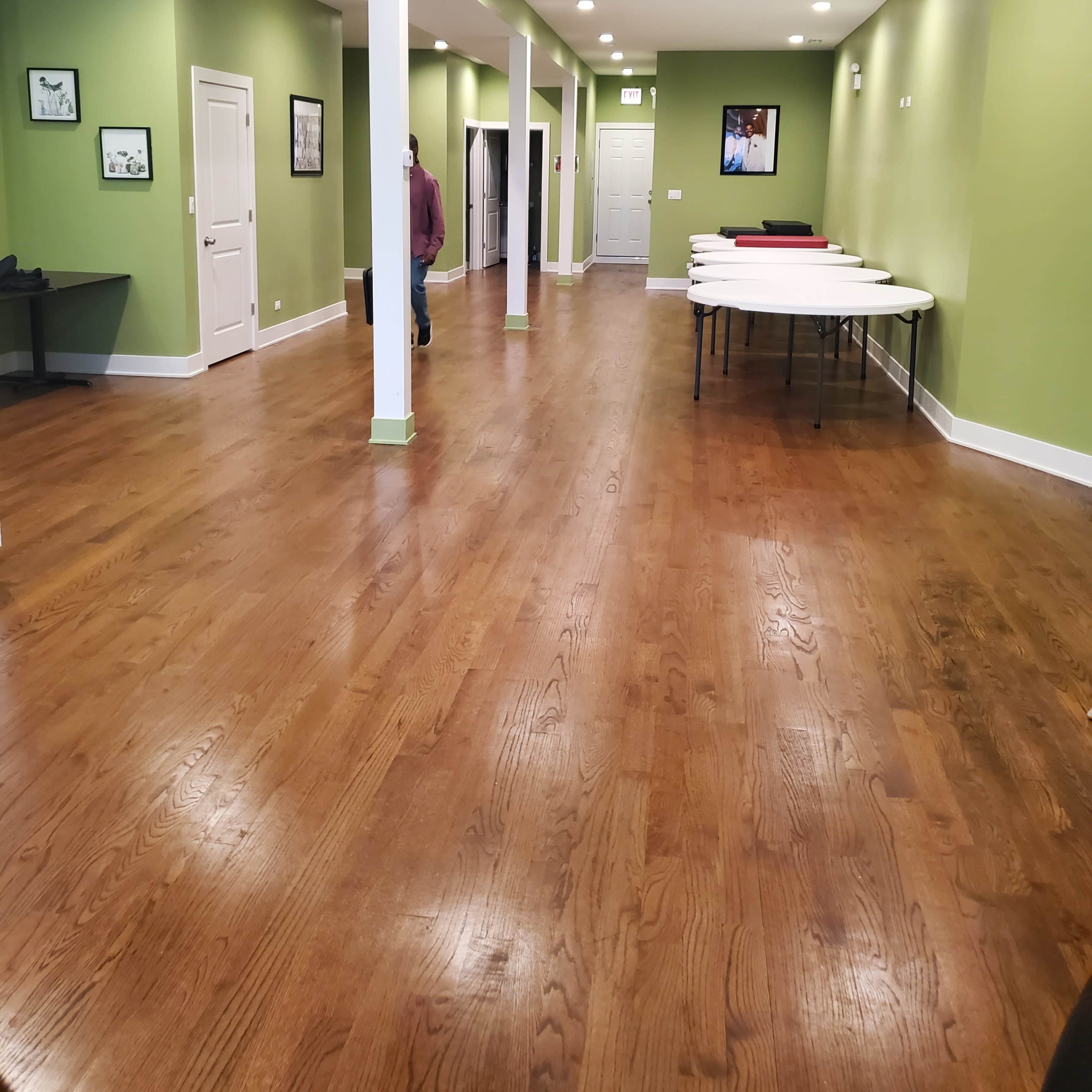 A spacious, green-walled room with polished wooden floors, featuring a row of tables along one side and doors leading to other areas.
