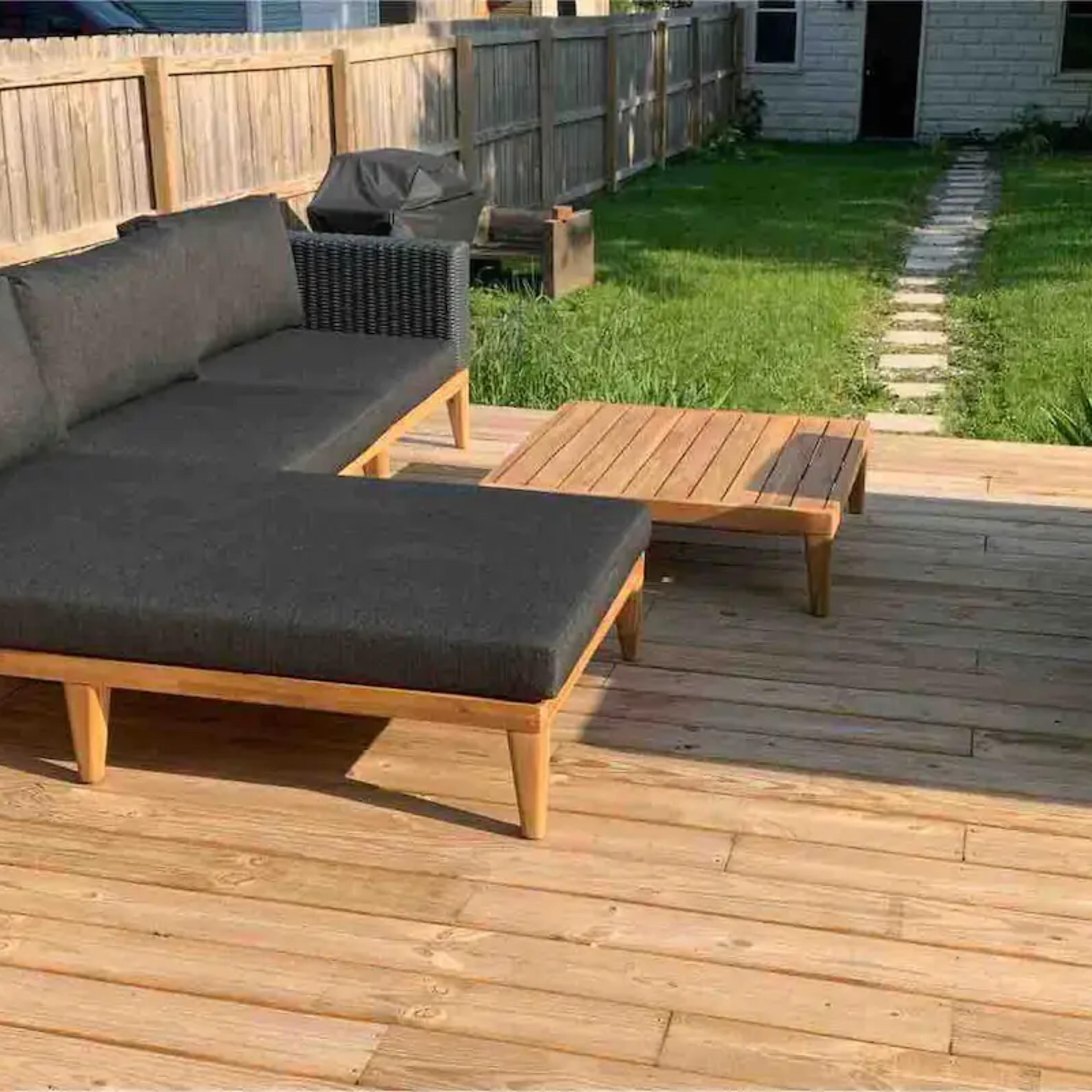 A wooden deck with a black cushioned sectional sofa and a wooden coffee table, surrounded by grass and a stone pathway in the background.
