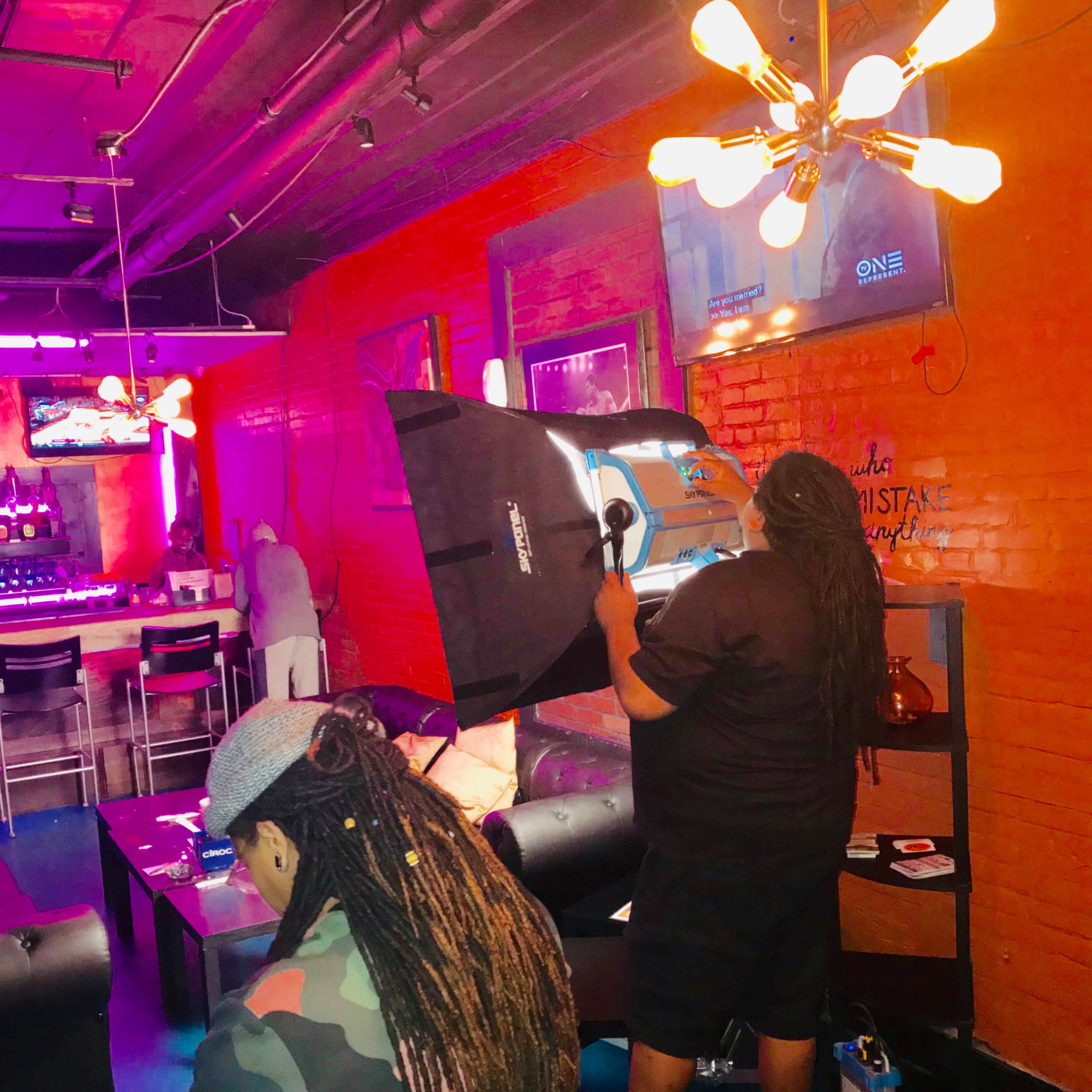 A person with long hair uses a light umbrella to set up a photo shoot in a lounge with red brick walls and colorful lighting.