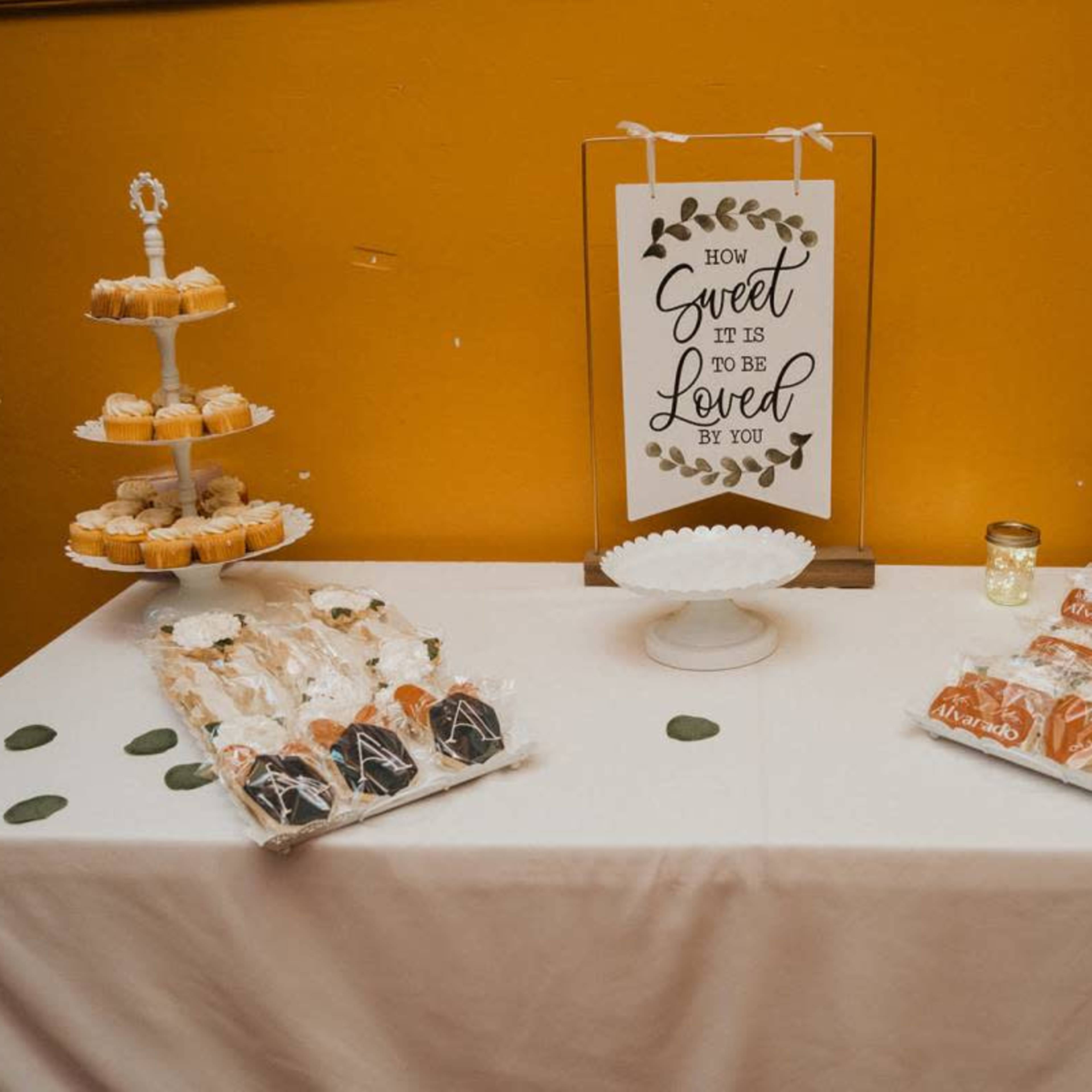 A dessert table features a sign that reads "How Sweet It Is to Be Loved by You," surrounded by various trays of cupcakes and decorated cookies.