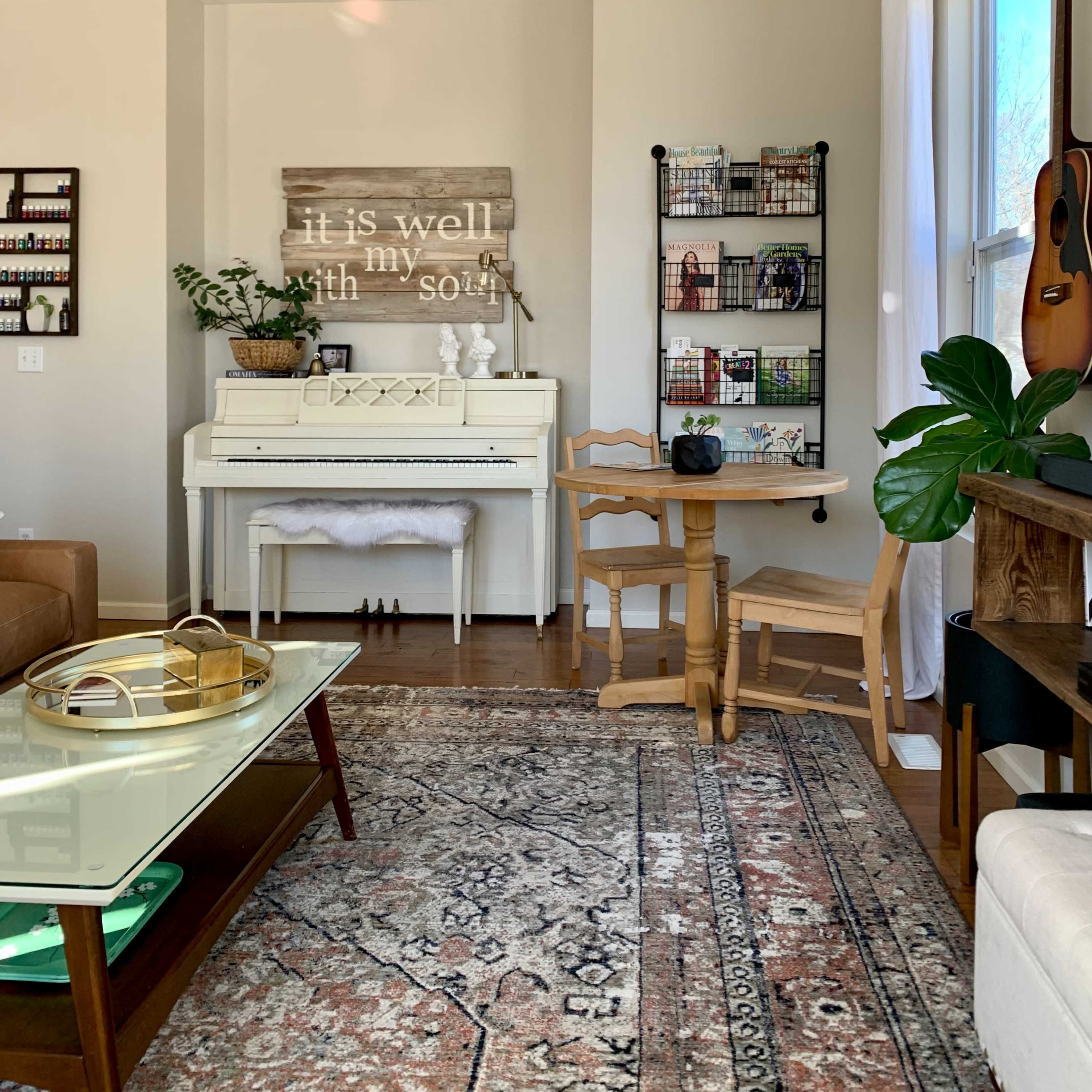 A cozy interior featuring a white piano, a small dining table, a bookshelf, and a decorative wall sign that reads "it is well with my soul."