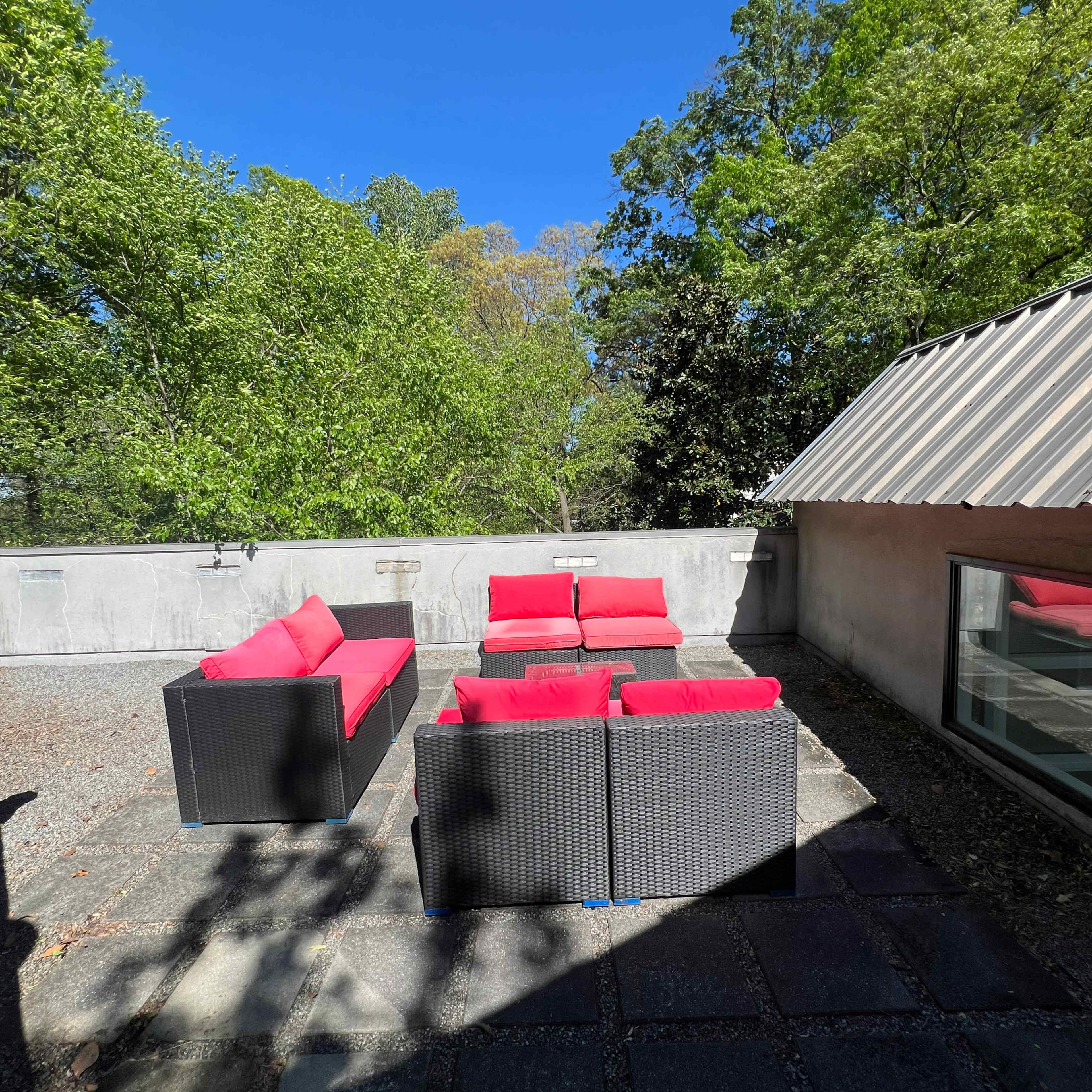 A patio area features a set of black wicker furniture with red cushions, surrounded by trees and clear blue sky.