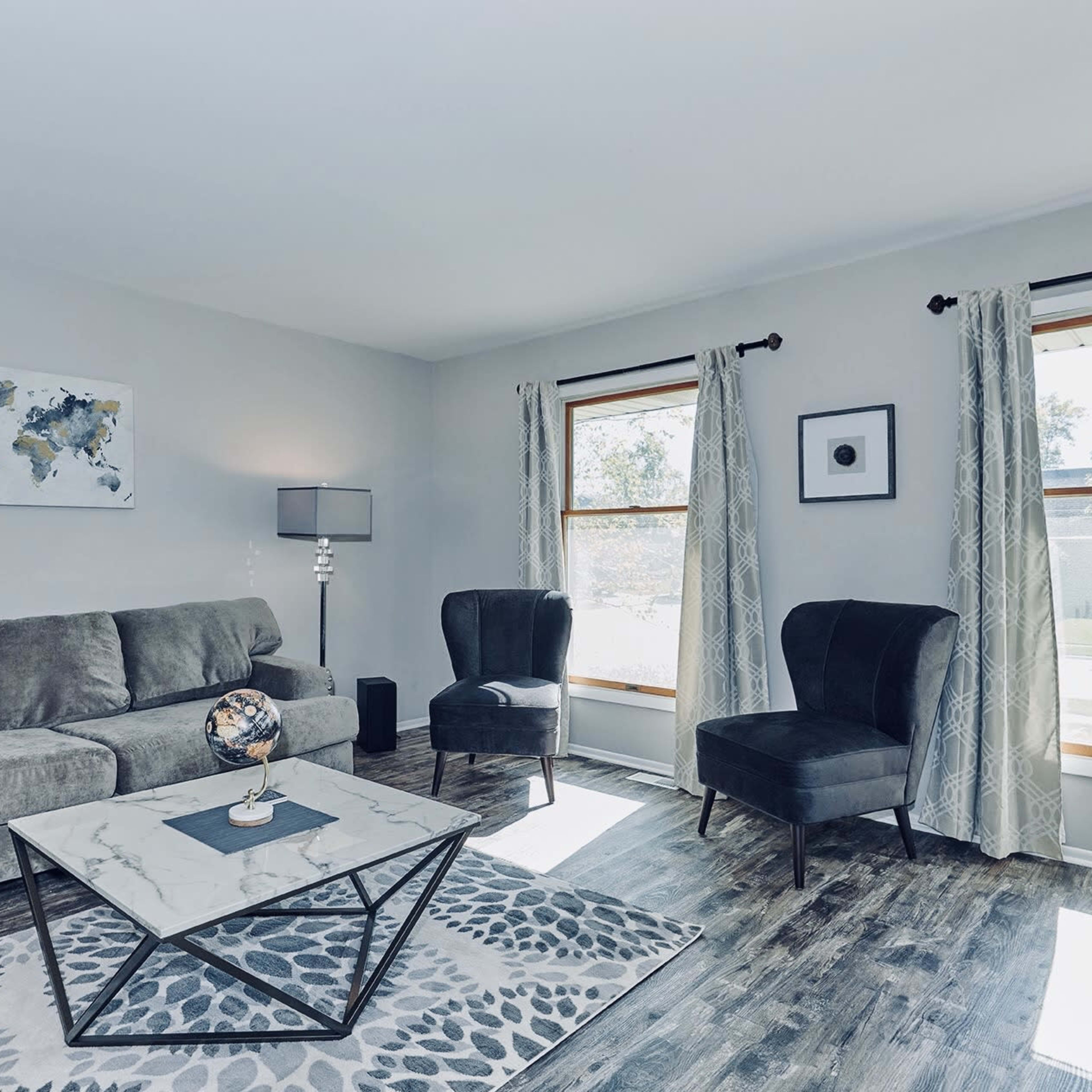 The living room features a gray sofa, two dark chairs, a marble coffee table, and large windows with light curtains.
