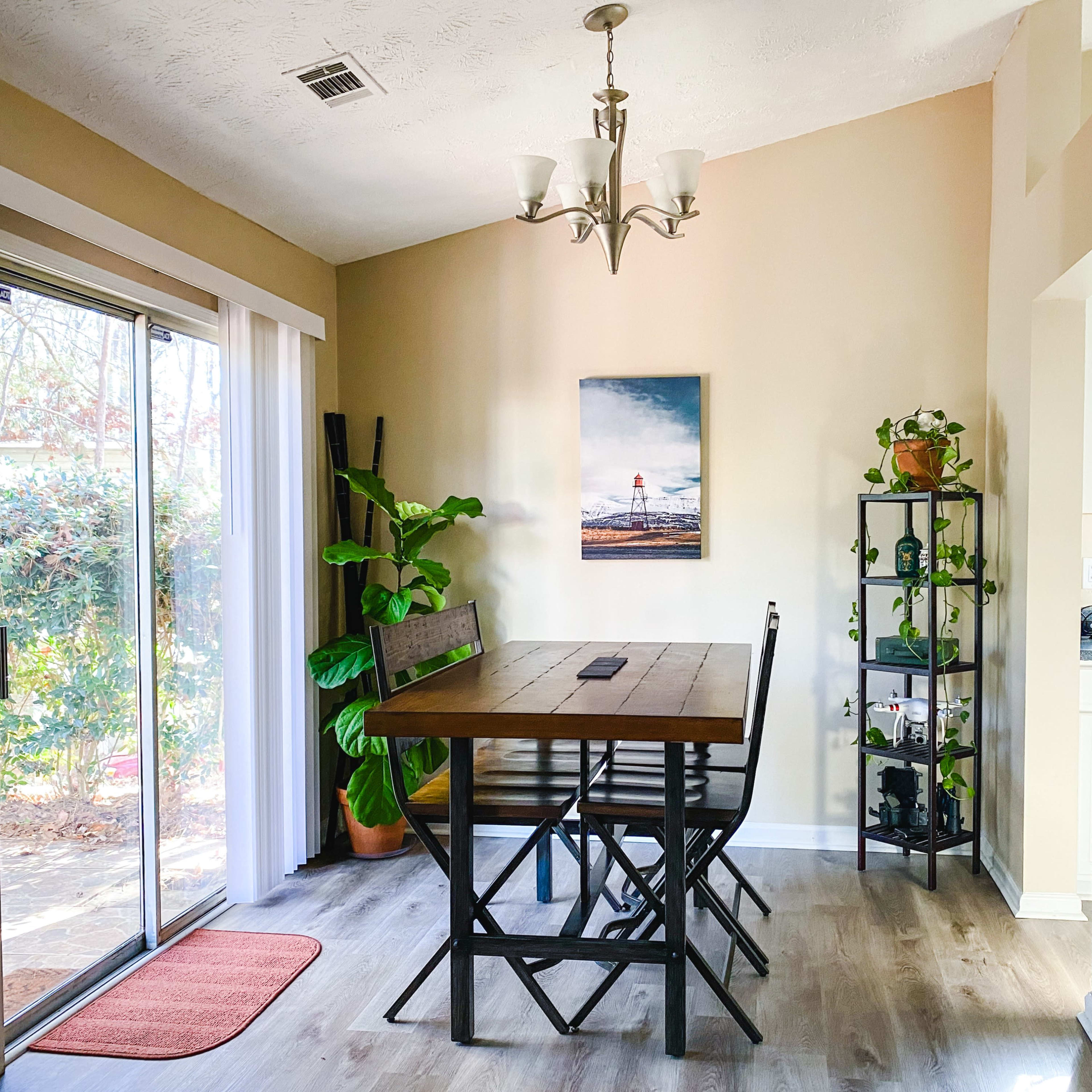 A dining area features a wooden table with four chairs, a large window with sliding glass doors, and a plant stand beside a wall-mounted art piece.