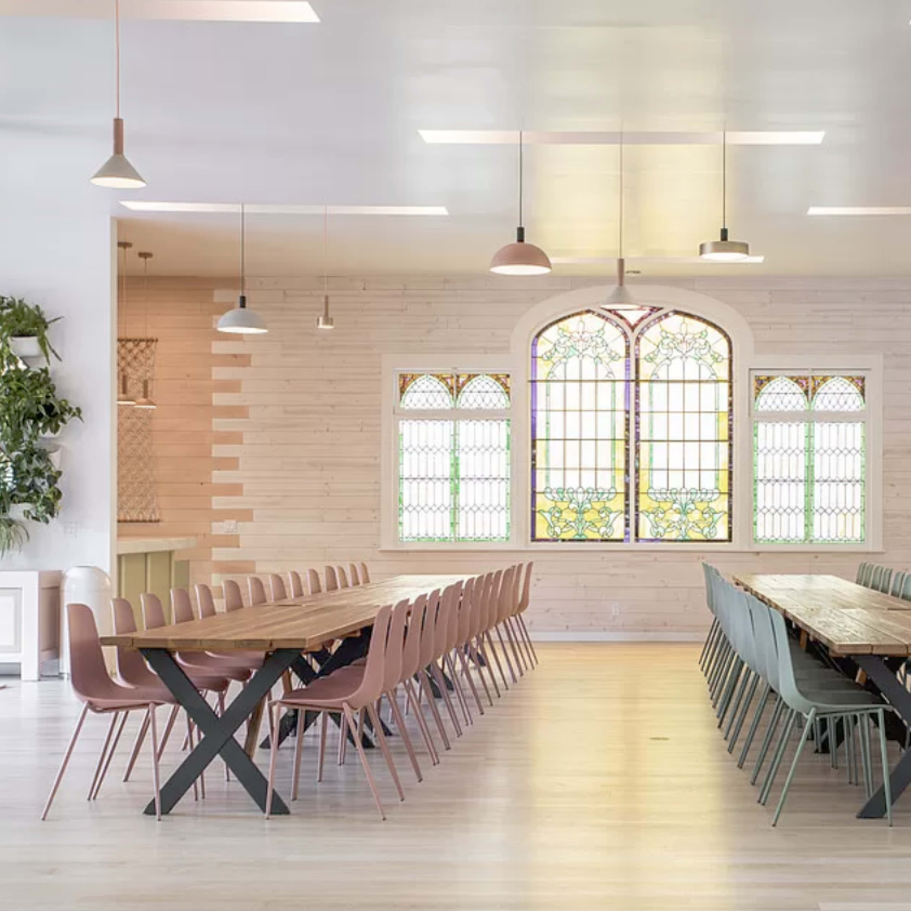 A spacious dining area with long wooden tables surrounded by modern chairs, featuring a large stained glass window on one wall.