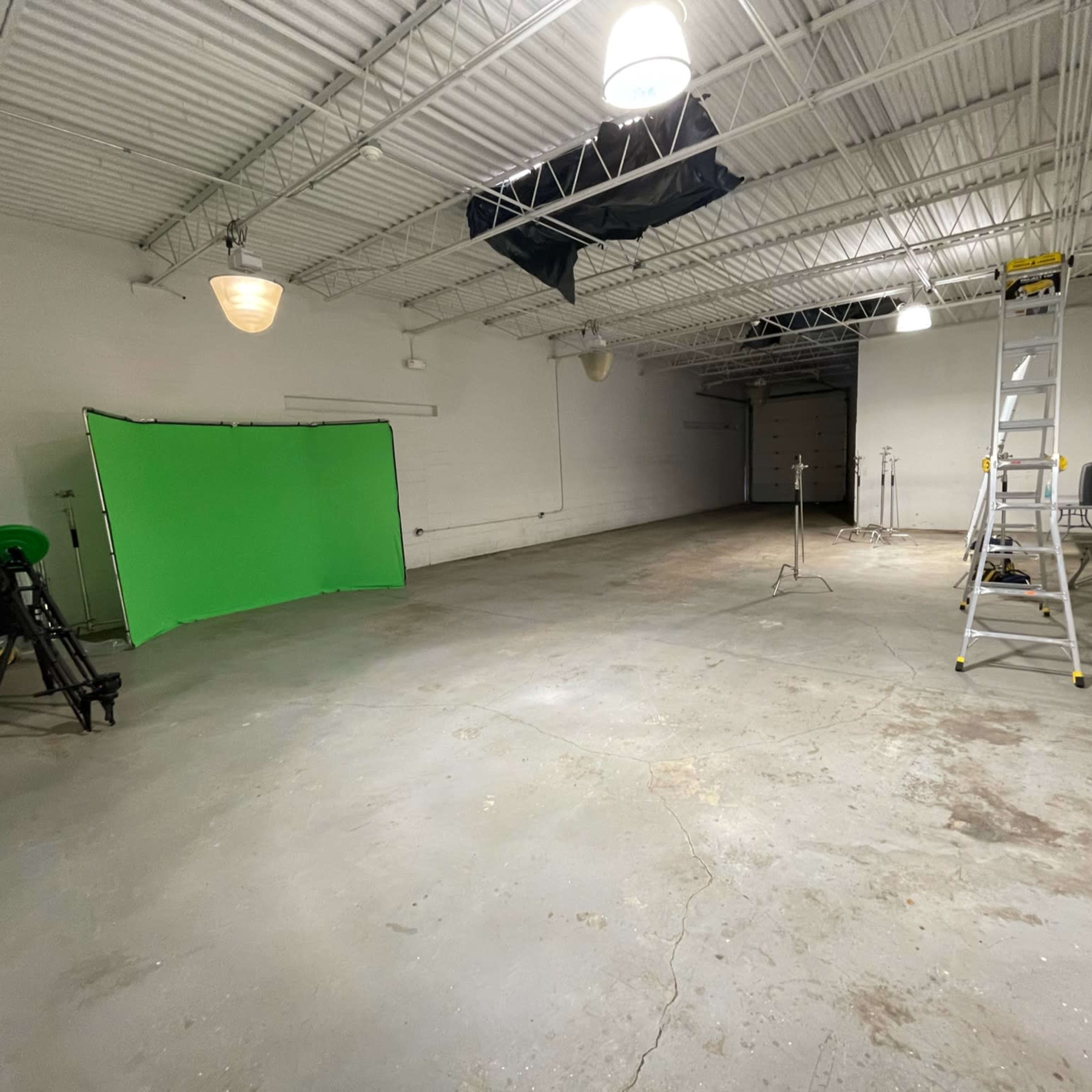 An empty warehouse space featuring a green screen setup, a ladder, and various lighting equipment.