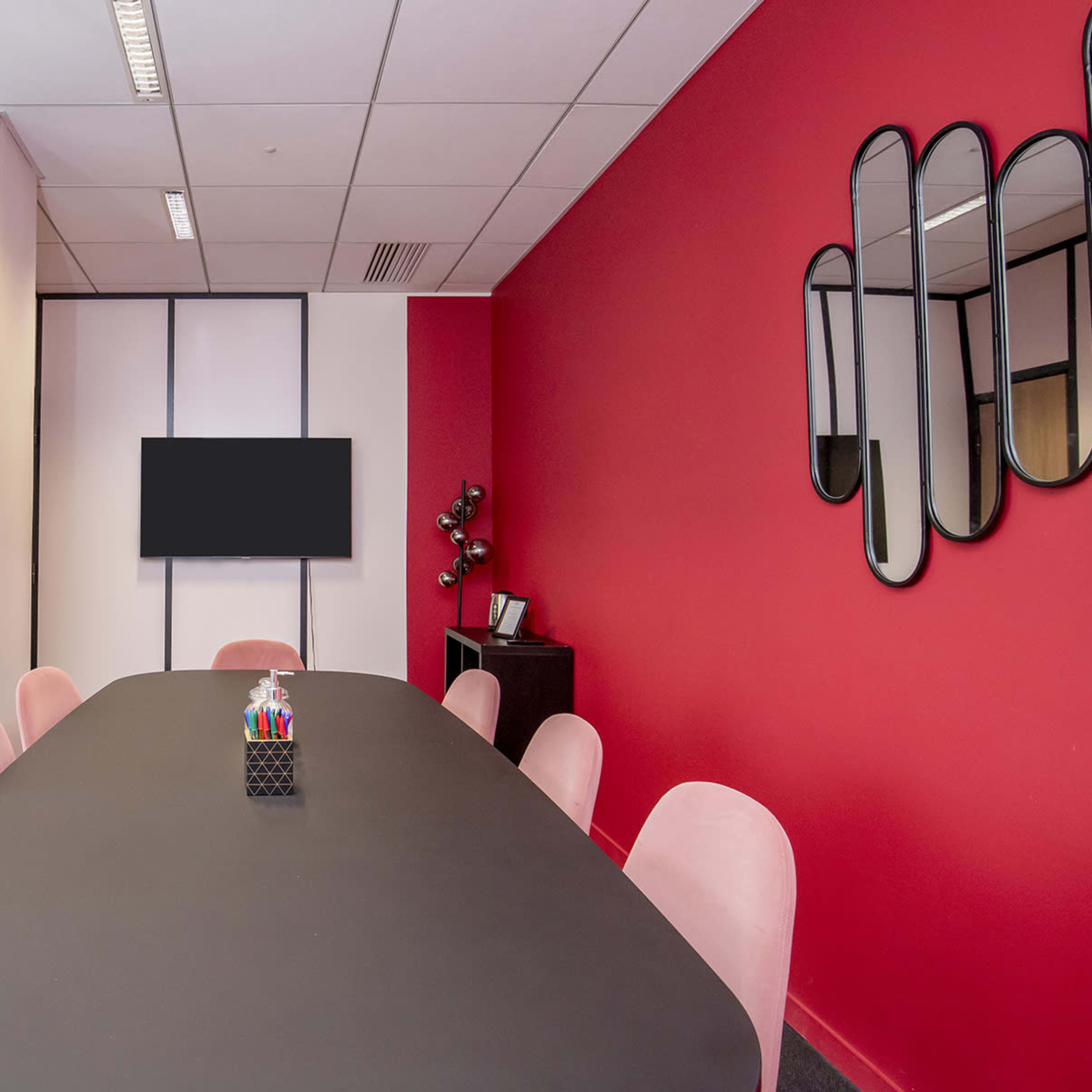 A modern conference room features a large black table surrounded by pink chairs, a wall-mounted screen, and decorative mirrors on the red wall.