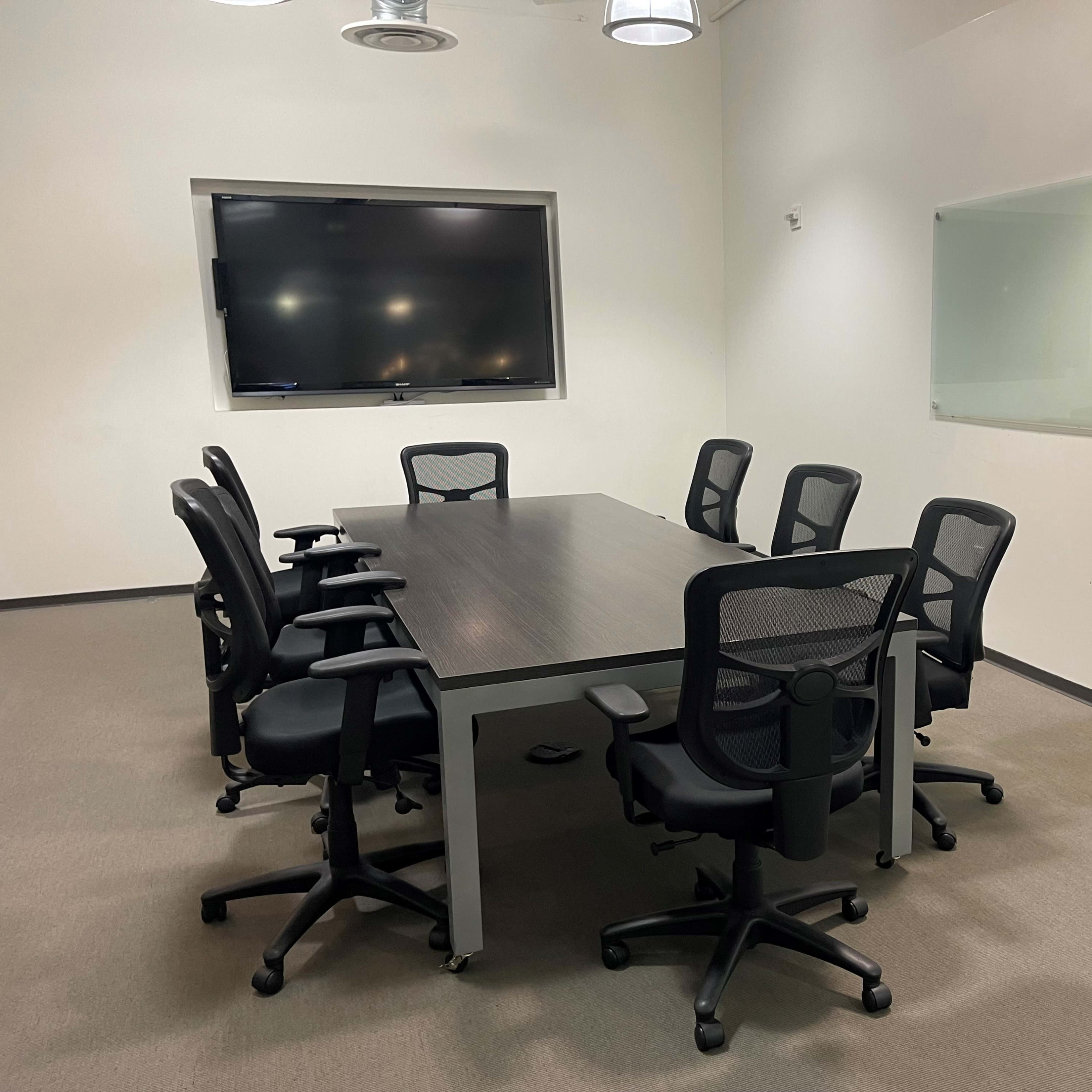 A conference room with a large table surrounded by eight black office chairs and a wall-mounted screen.