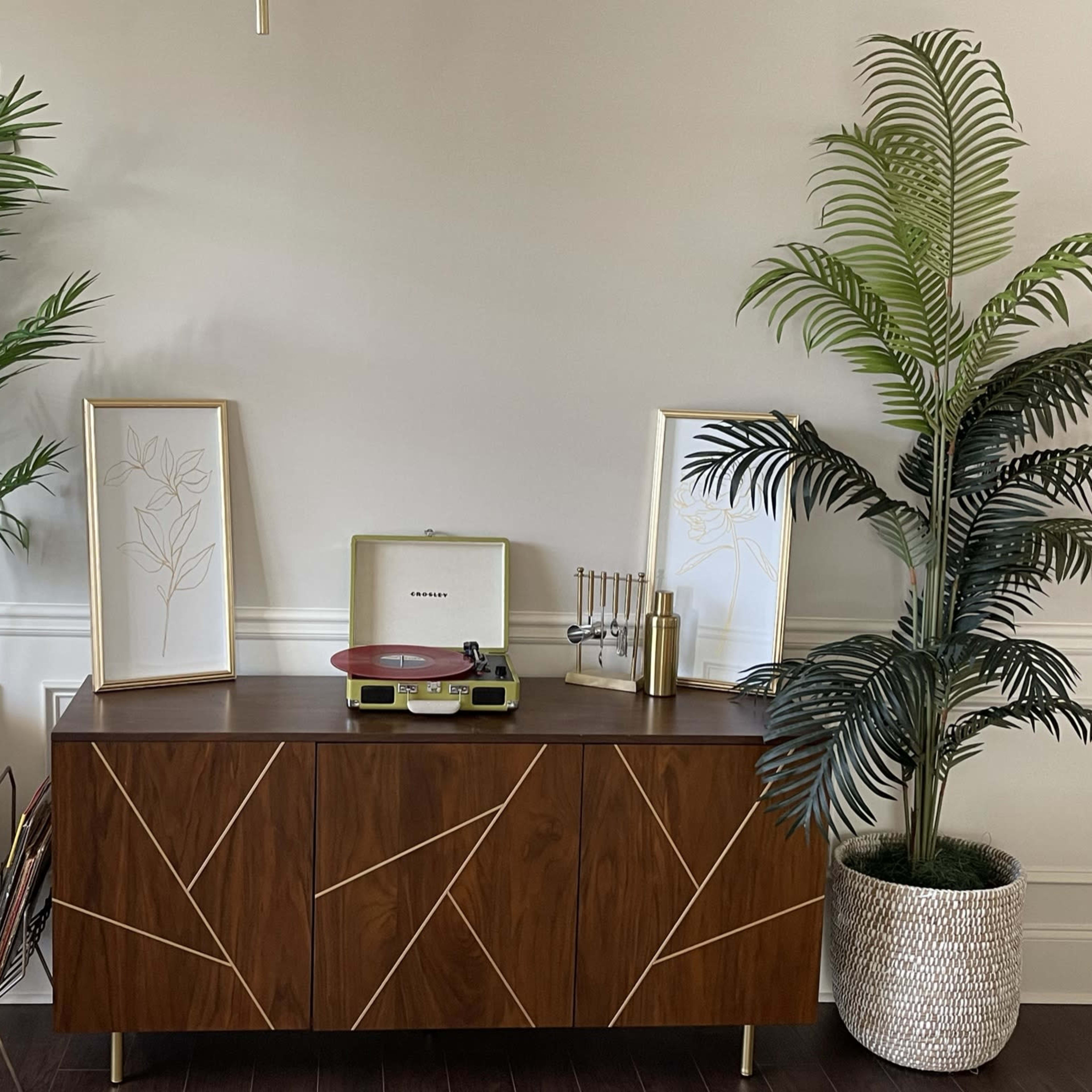 A wooden sideboard with a record player sits between two potted plants, flanked by framed botanical art on the wall.