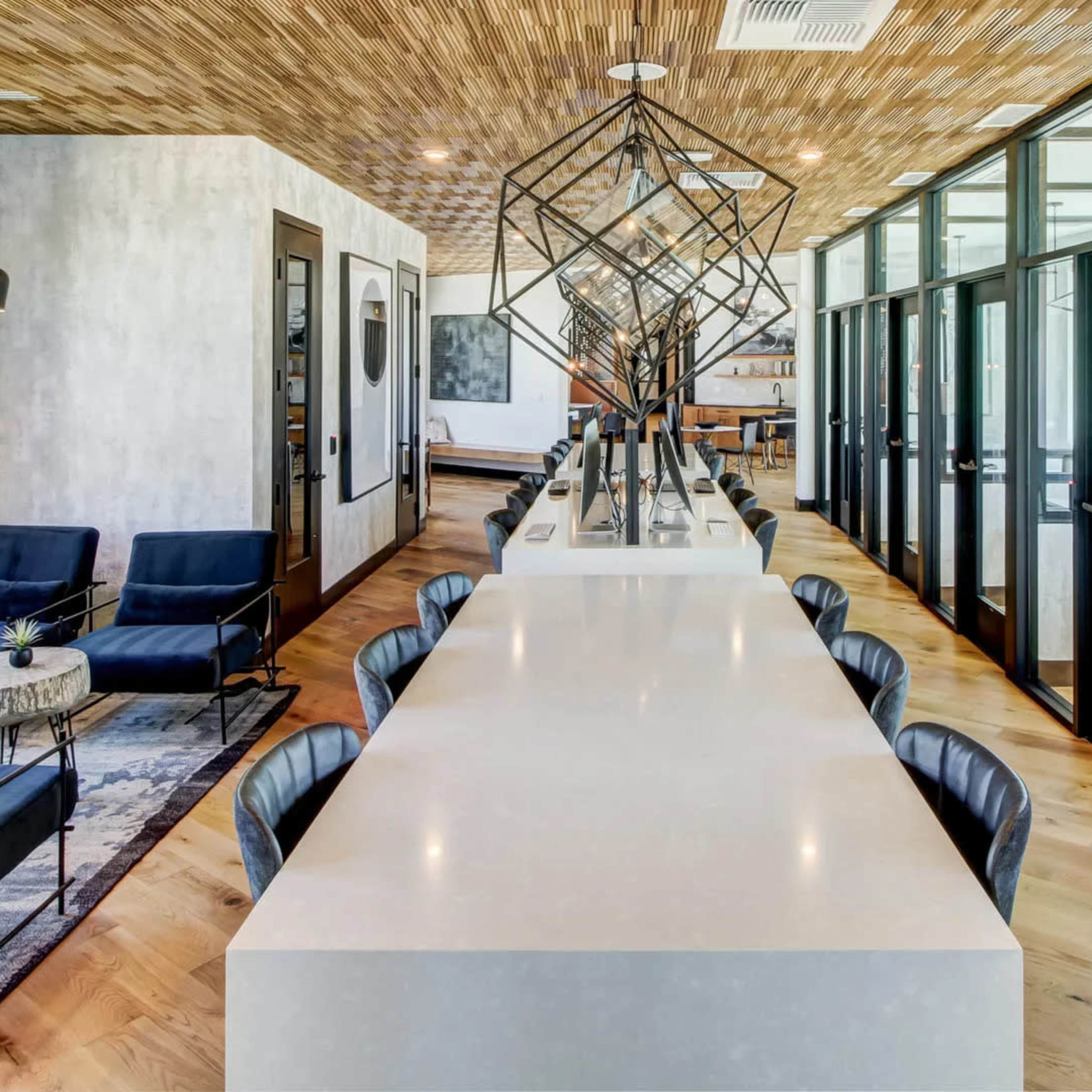 A modern office space featuring a long white table with black chairs, surrounded by seating areas with blue upholstered chairs and decorative tables, under a wooden ceiling and large windows.