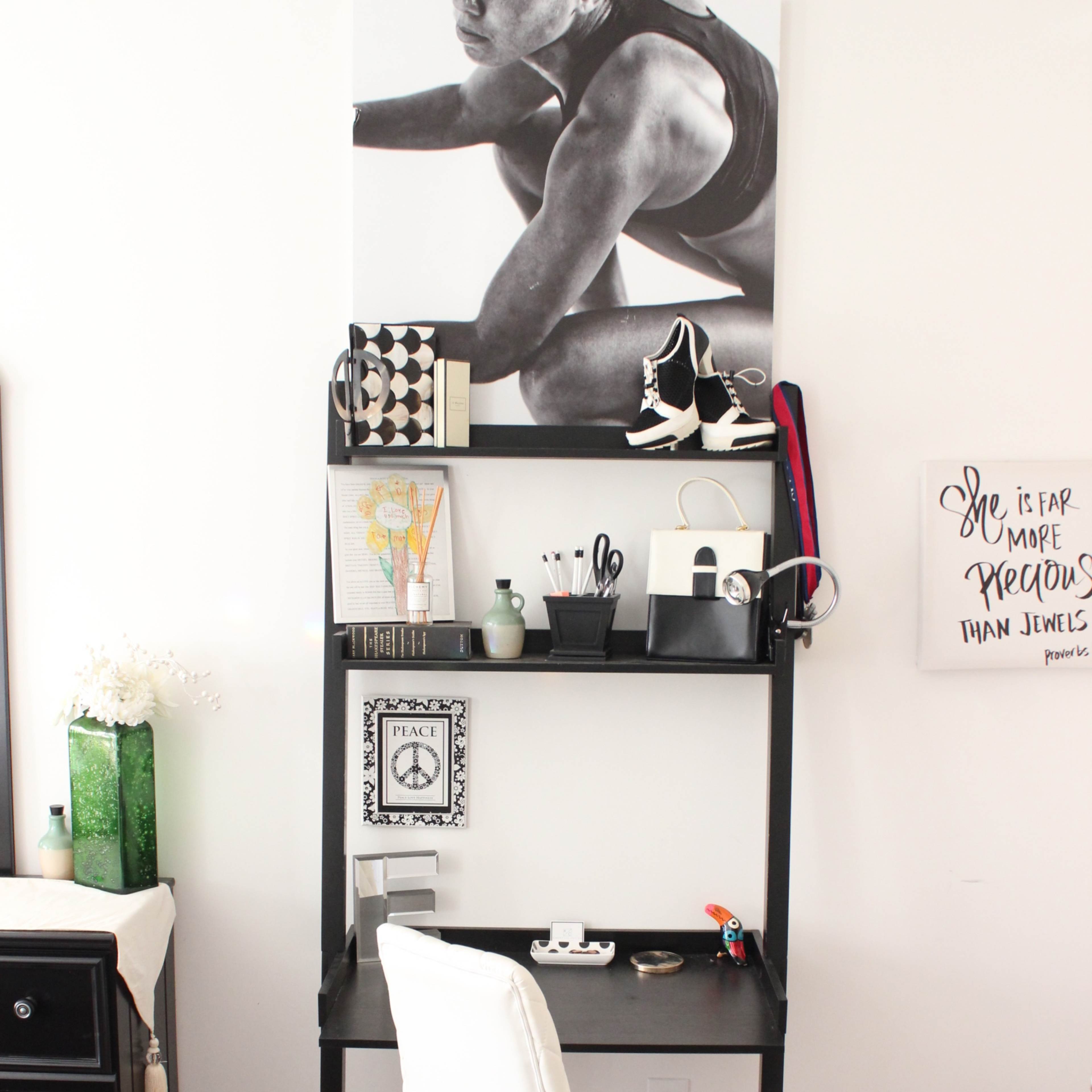 A minimalist desk setup features a black desk with a rolling chair, shelves displaying various decorative items and office supplies, and a large black-and-white poster on the wall above.