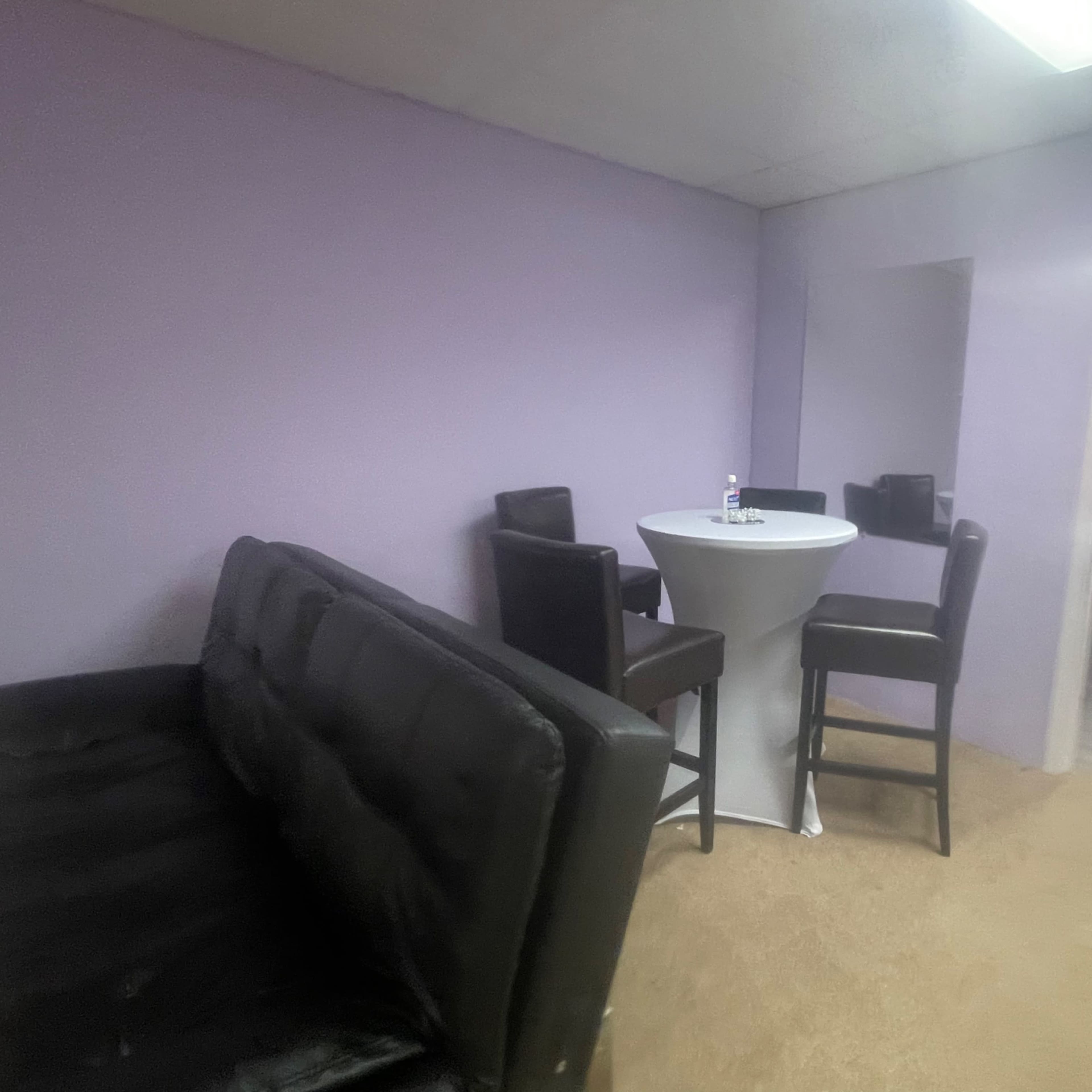 A small room with purple walls, featuring a round table surrounded by tall chairs, a black couch, and an adjoining room with a table visible.
