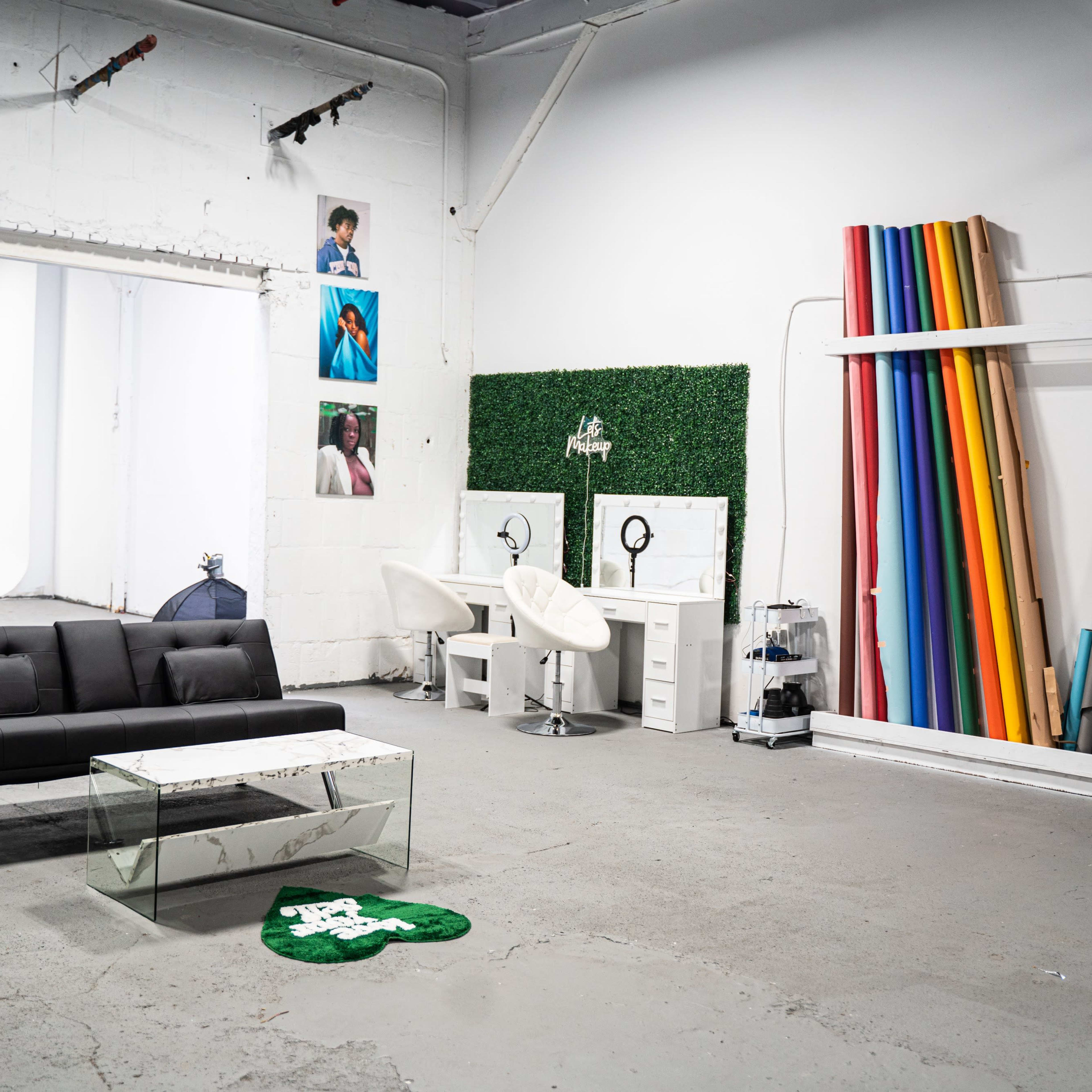 An industrial-style studio space featuring a black sofa, a glass coffee table, a two-mirror vanity station, and a collection of colorful backdrop rolls on the wall.