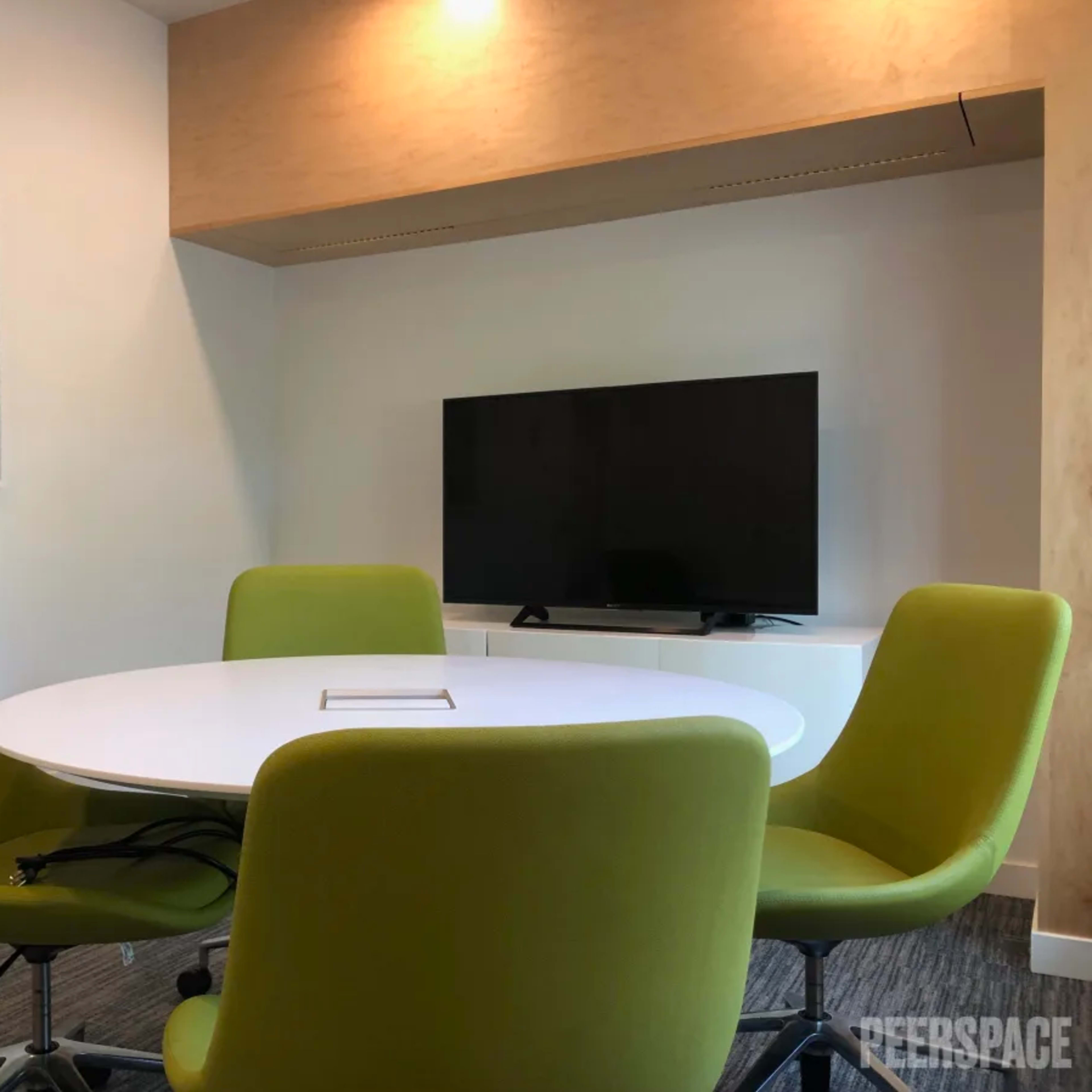 A round white table with four green chairs is set in a brightly lit conference room featuring a wall-mounted television and an abstract painting.