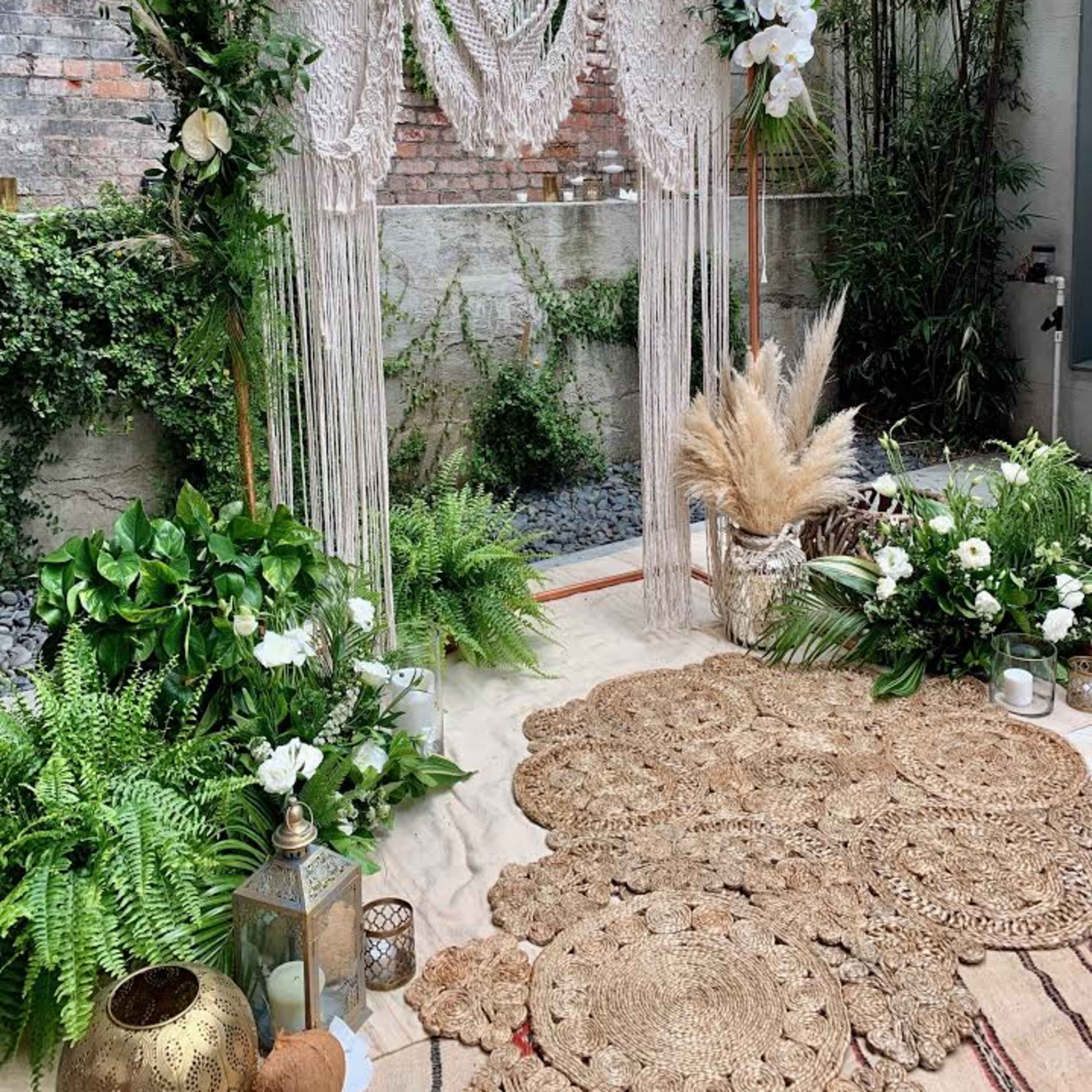 A decorated outdoor space featuring a macramé backdrop adorned with white flowers, surrounded by various greenery and natural decor elements on a sandy floor.